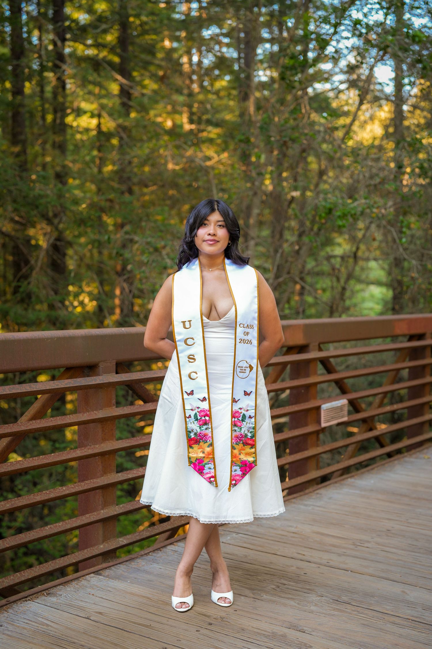 Graduate wearing a white dress and floral UCSC stole stands on a wooden forest bridge beneath sunlit trees.
