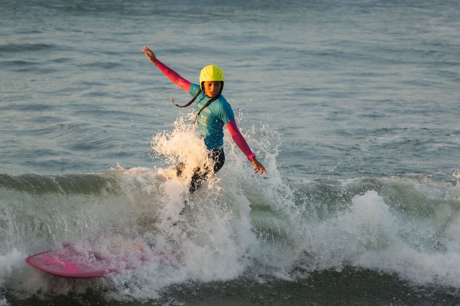 Surfer in a yellow helmet and teal wetsuit rides a longboard through splashing white water with arms outstretched.