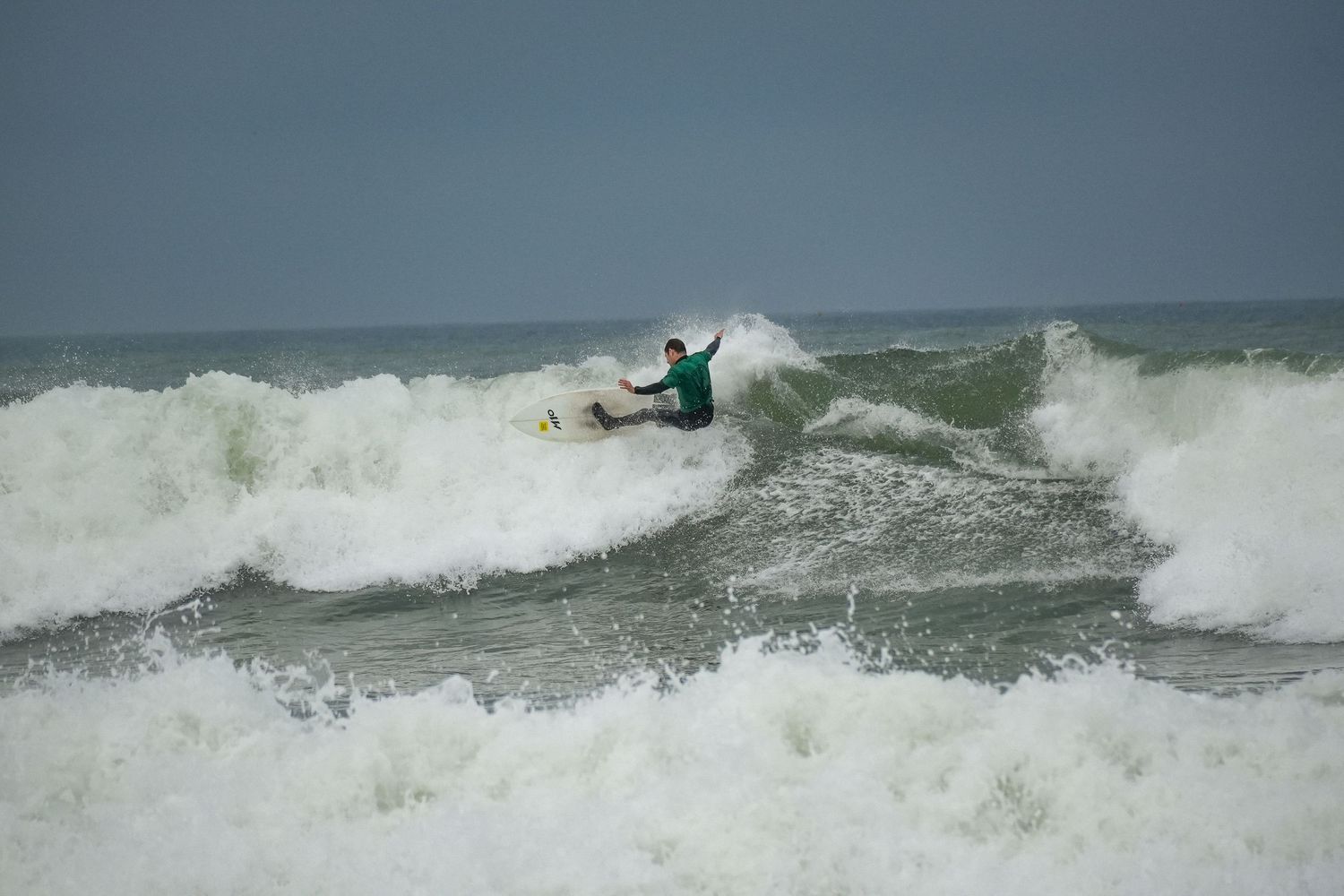 Surfer in a green rash guard rides a powerful breaking wave surrounded by whitewater under an overcast sky.