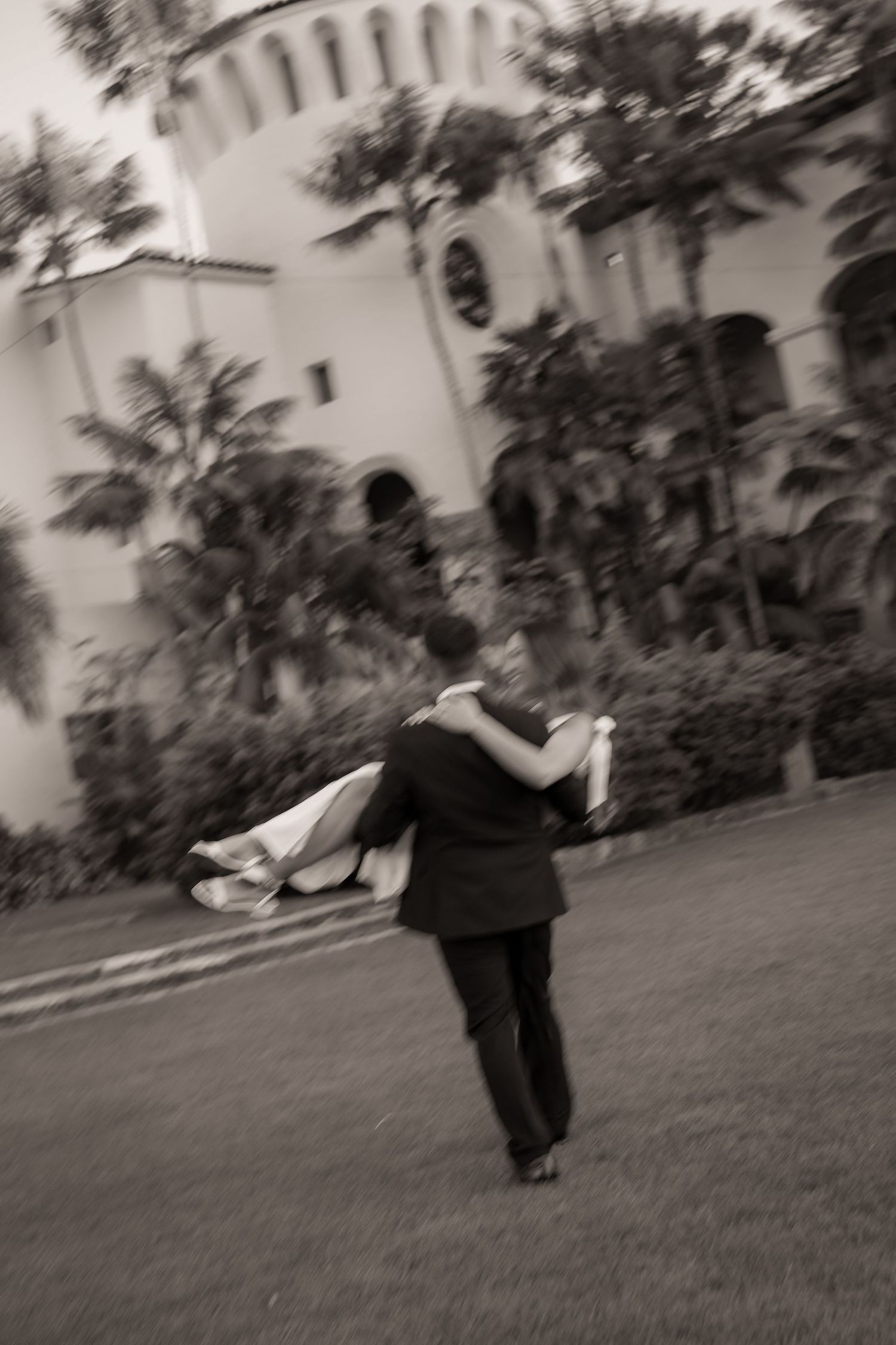 Blurred black and white photo of a groom carrying his bride across a lawn toward a Spanish-style building framed by palm trees.