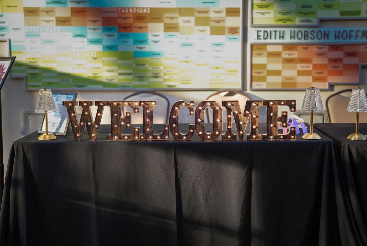Lighted marquee letters spell WELCOME on a black draped table in front of a colorful wall chart at a museum event.