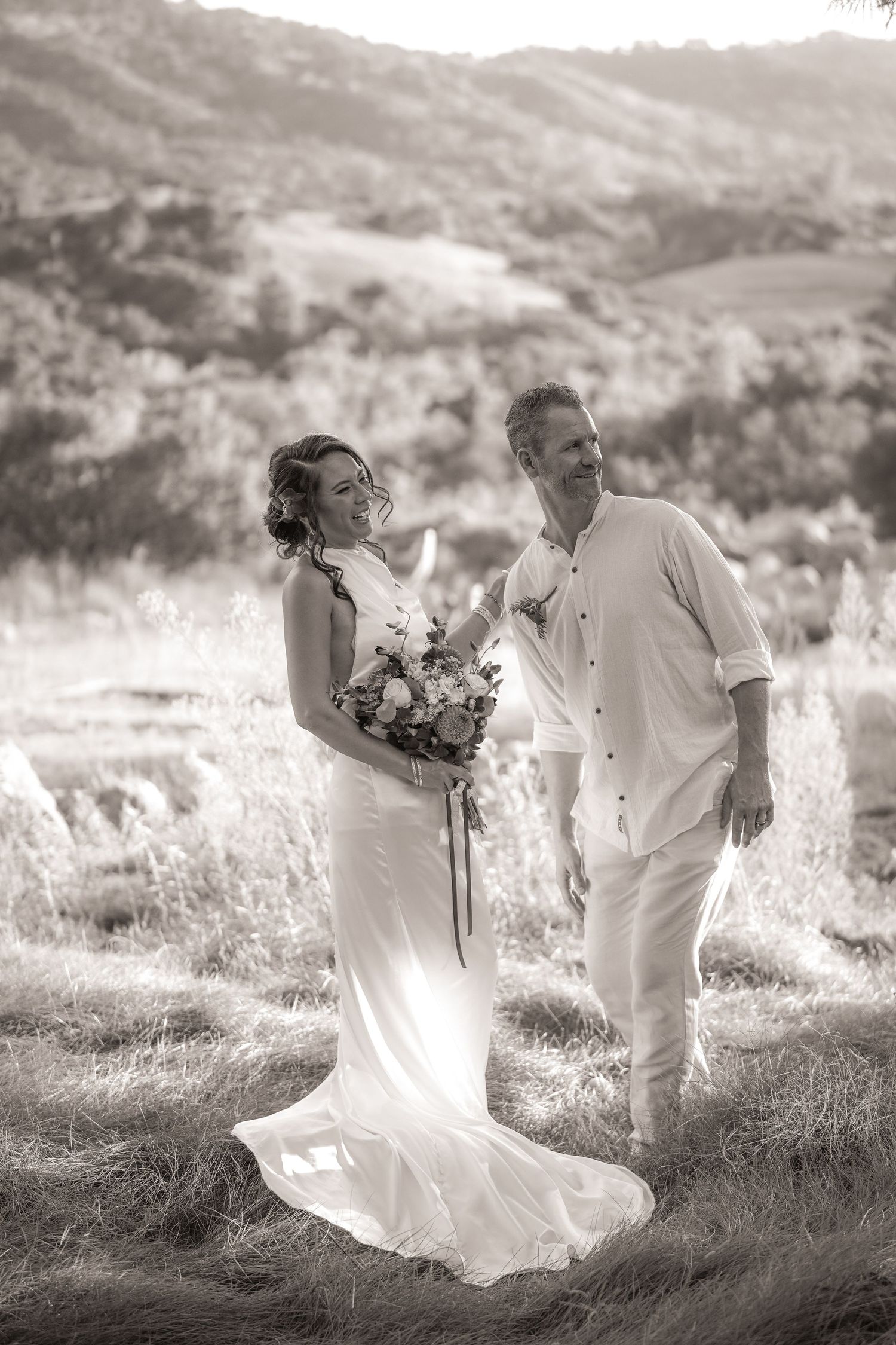Black and white photo of a smiling bride and groom standing in a grassy field with rolling hills behind them.