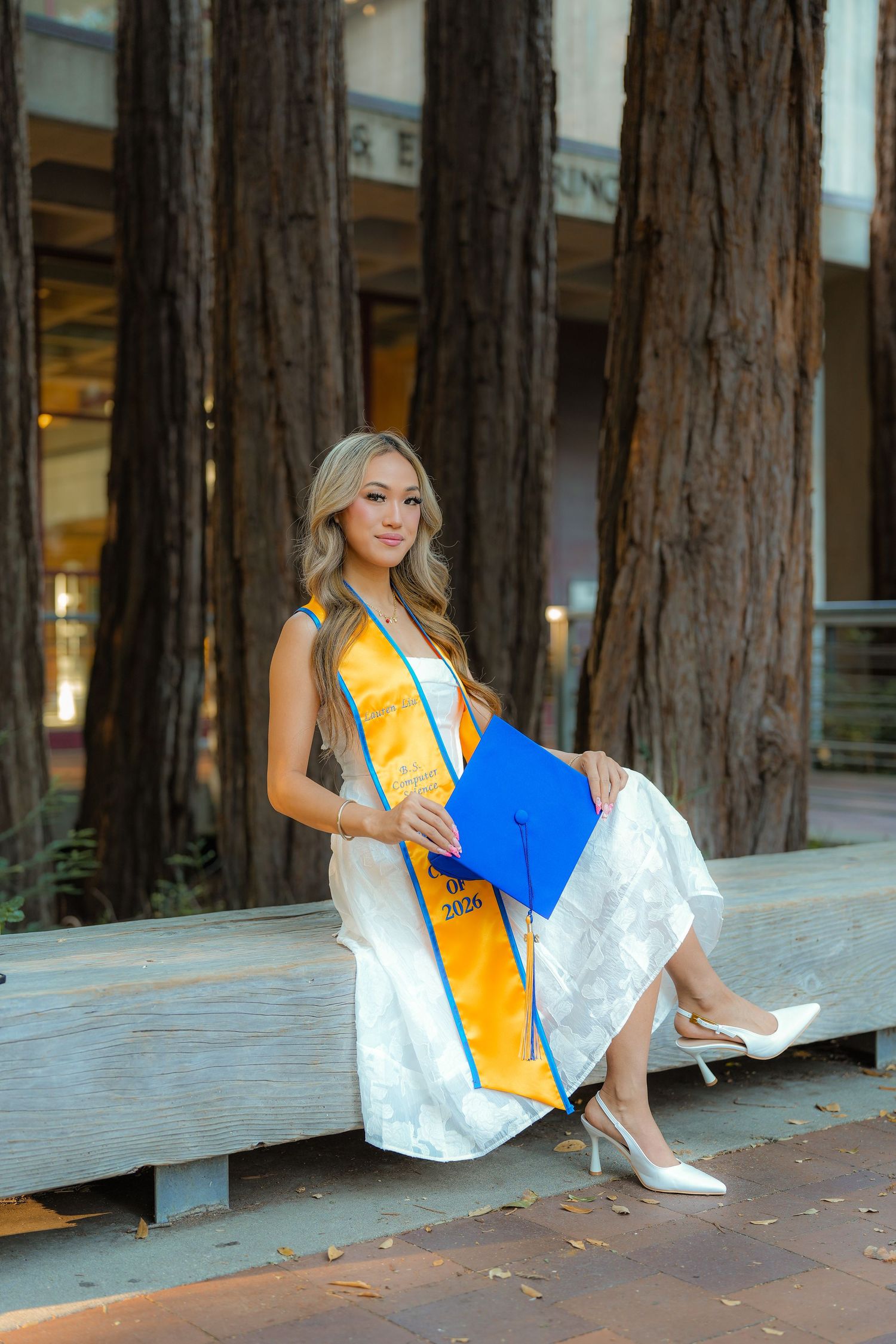 Graduate in a white dress and gold stole sits on a stone bench among tall redwood trees, holding a blue cap and wearing white heels.