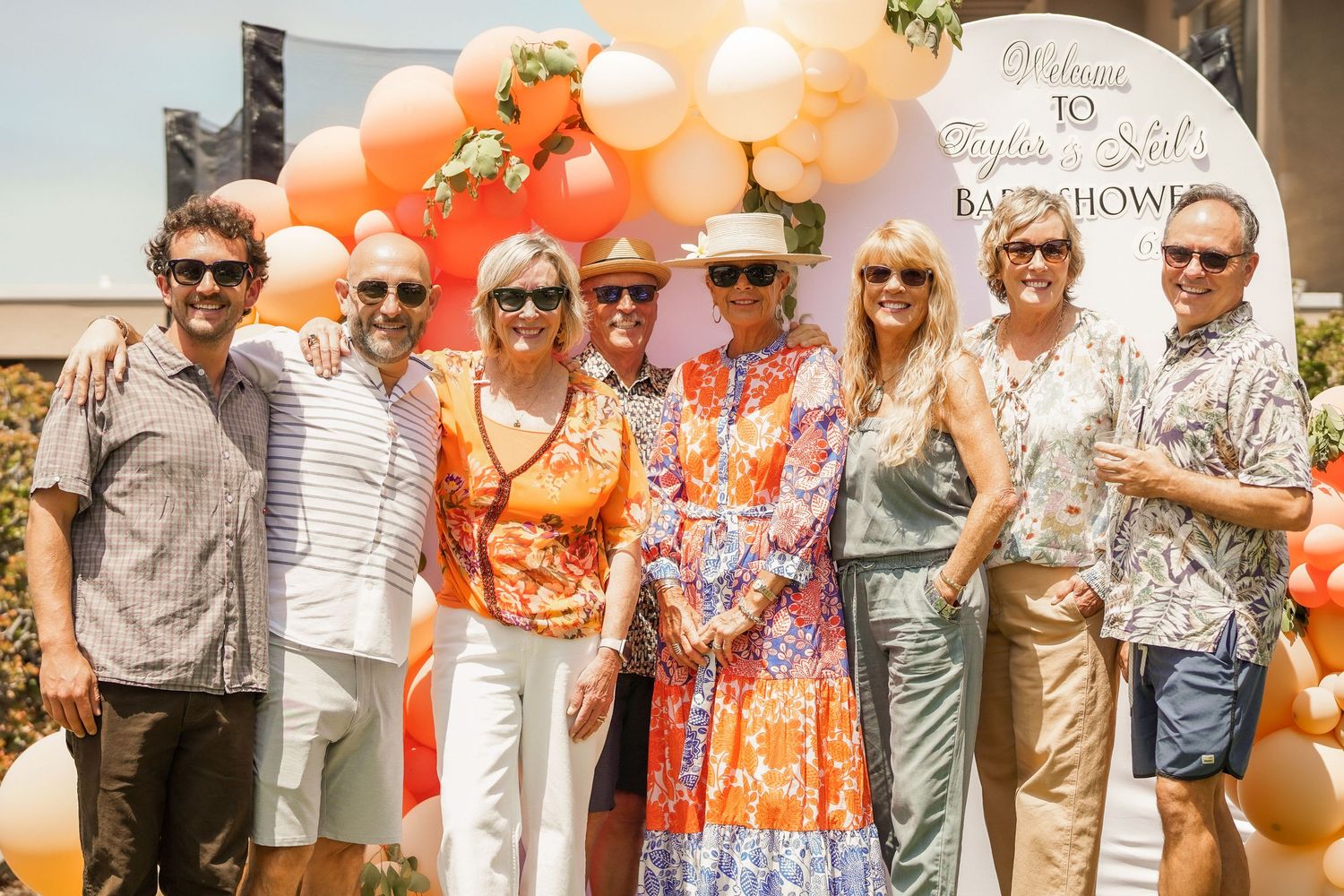 Group of smiling friends in colorful summer outfits and sunglasses pose together at an outdoor party with orange and peach balloons.