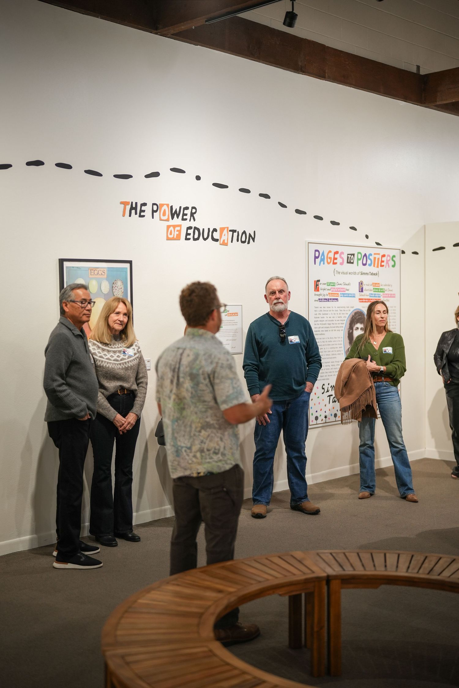 Visitors gather in a gallery beneath a The Power of Education banner while a man in a floral shirt addresses the group.
