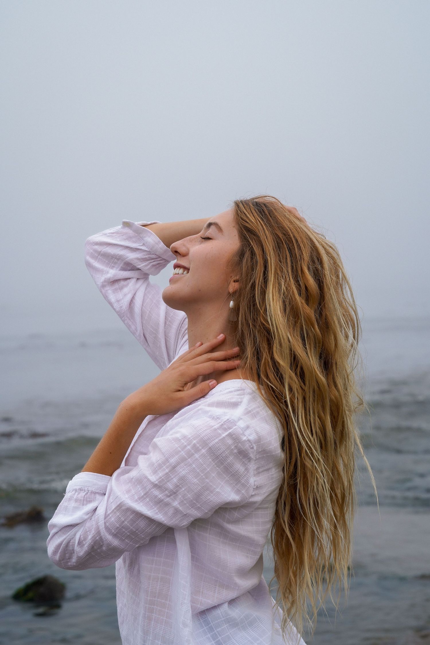 Woman in a white linen shirt laughs with her head tilted back and hand in her long wavy hair beside a foggy ocean.