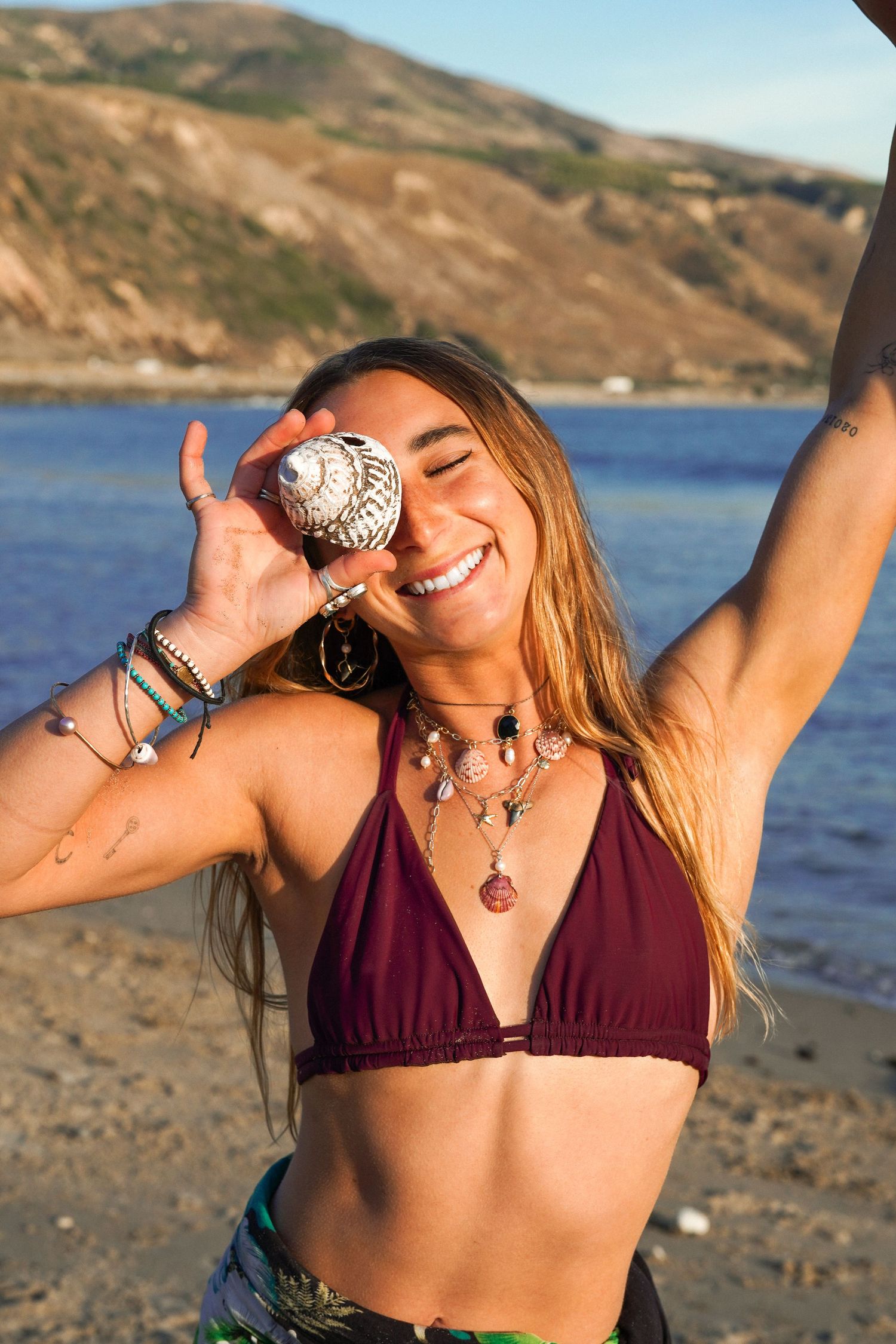 Laughing woman in a maroon bikini top holds a seashell to her cheek, arm raised, on a golden-hour beach by the ocean.