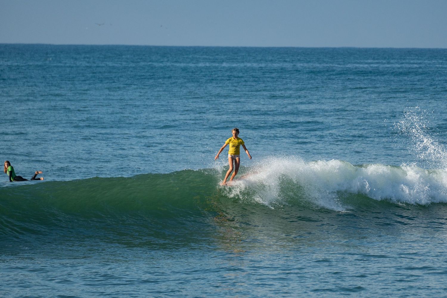 Surfer in a yellow top glides across the face of a breaking wave on the open blue ocean.