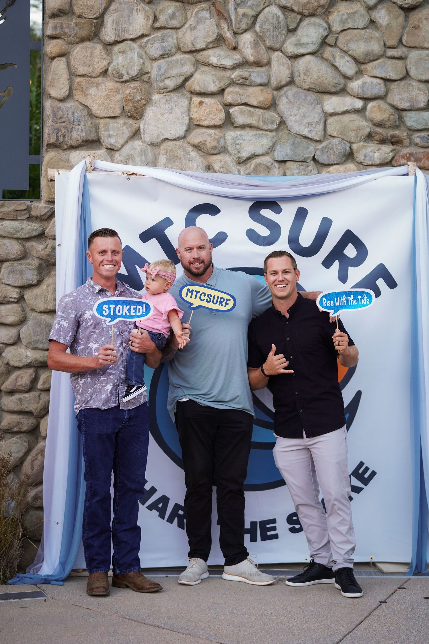 Two men and a toddler hold photo-booth signs while posing in front of an MTC Surf backdrop with a stone wall behind them.