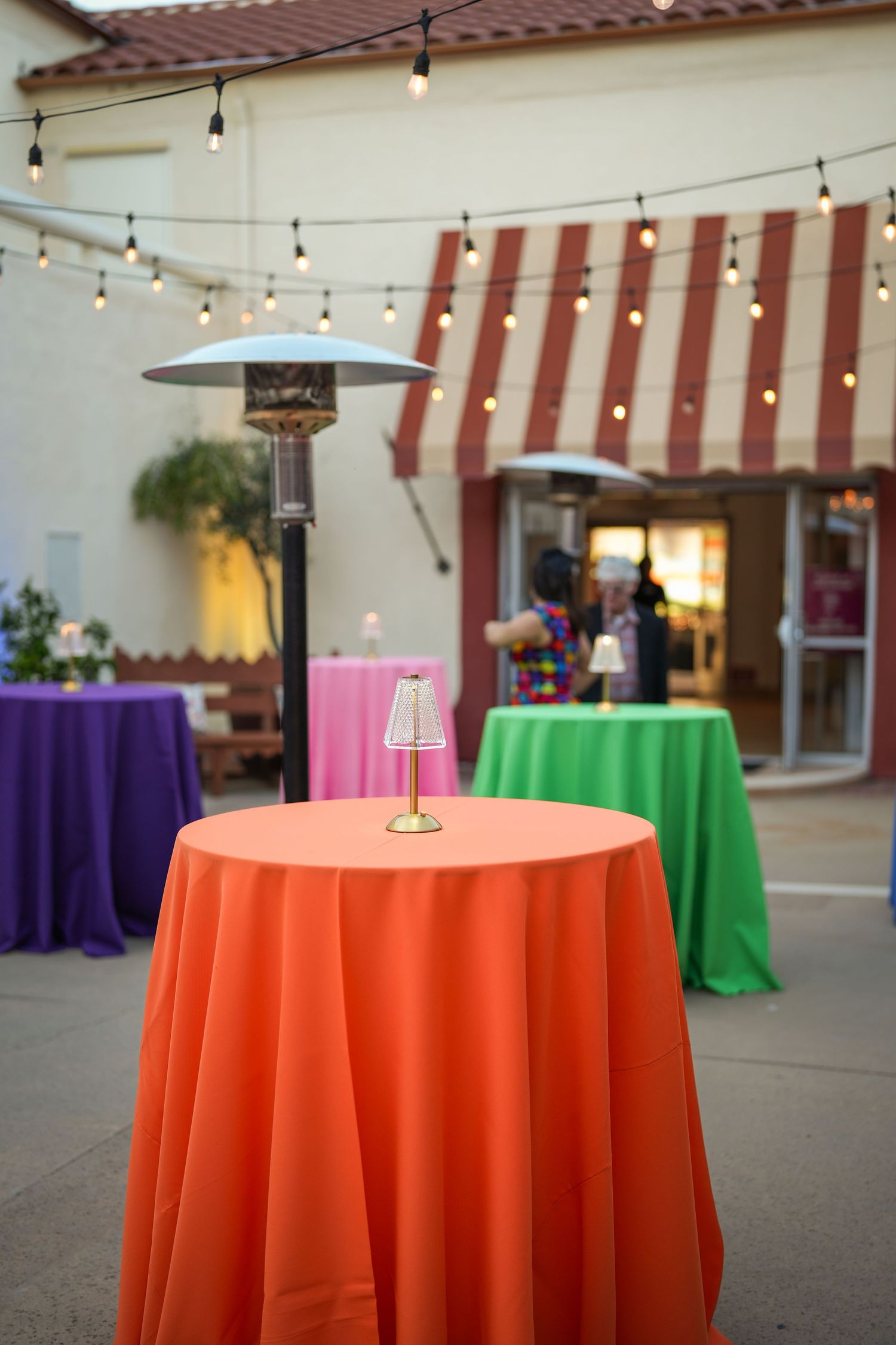 Orange, green and purple draped cocktail tables with gold lamps under string lights and patio heaters in an outdoor courtyard.
