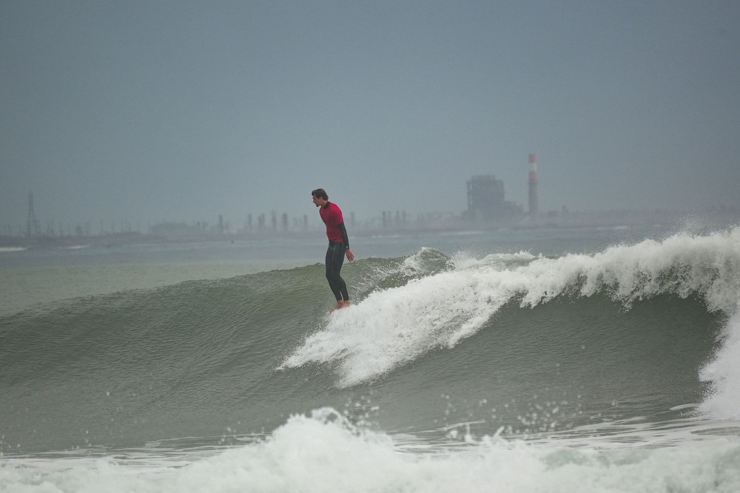 Longboard surfer in a red shirt stands tall on a rolling wave with a hazy coastal skyline behind.