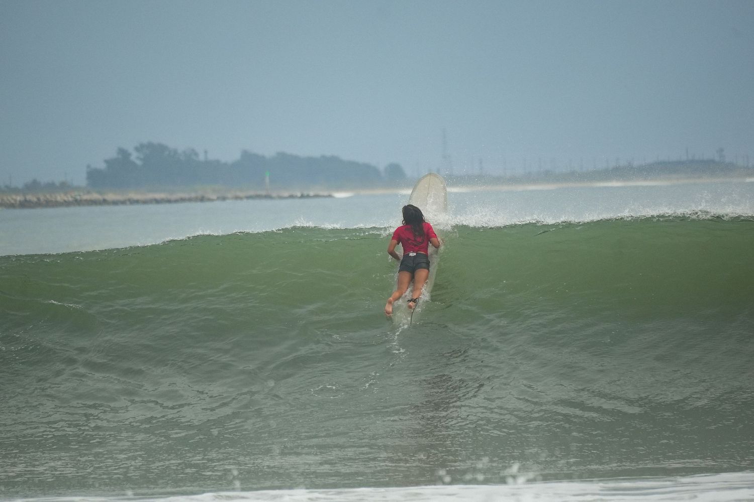 Surfer in a red top rides a smooth green wave toward the shore under a hazy gray sky.