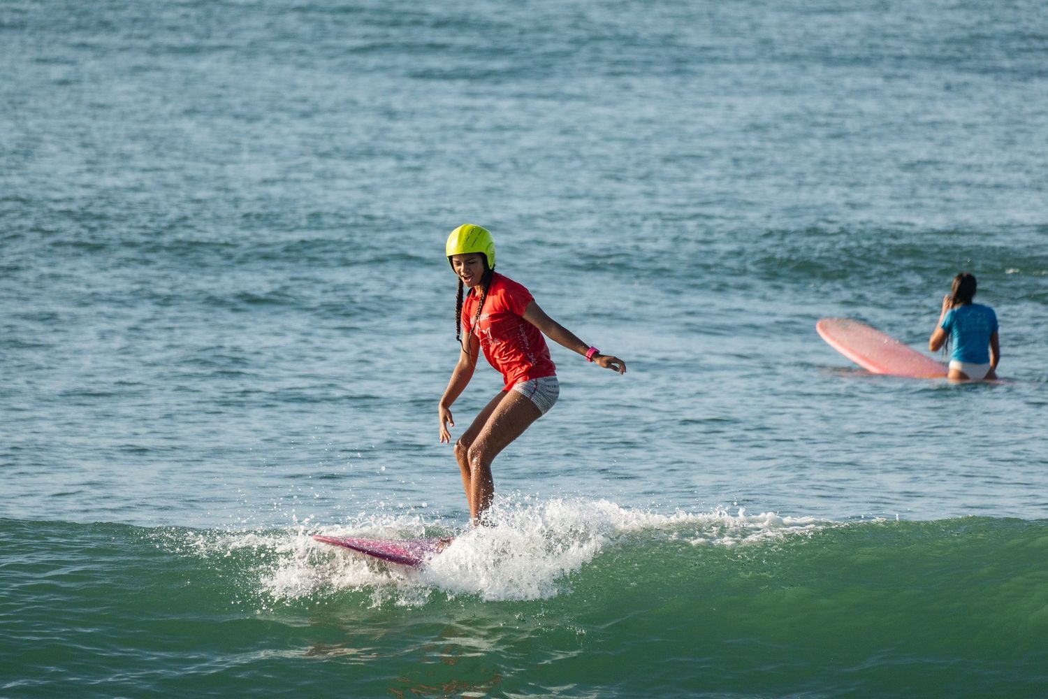 Surfer wearing a lime green helmet and red shirt balances on a longboard riding a small green wave.