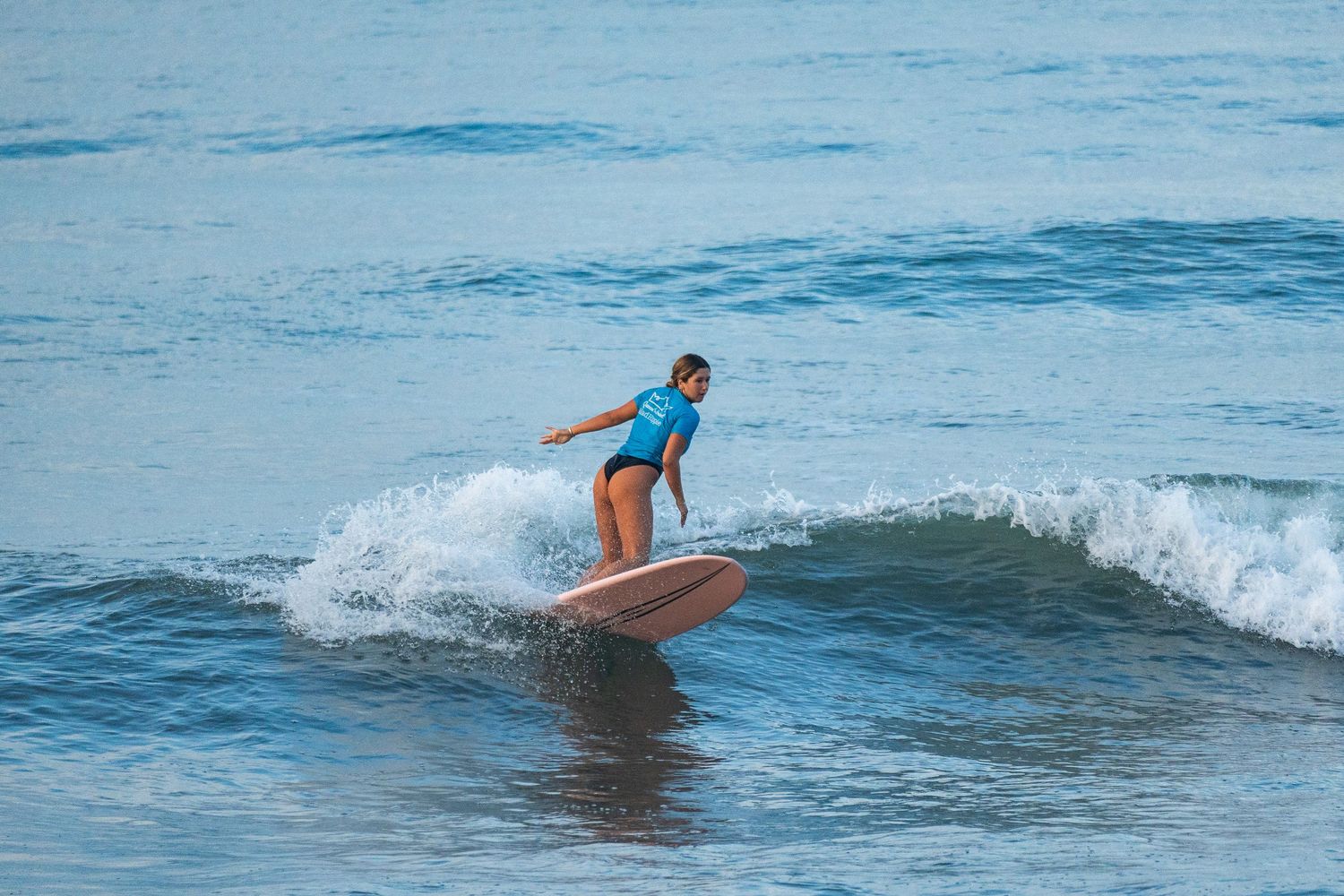 Surfer in a blue top rides a small wave on a longboard with arms out for balance in calm blue-green water.