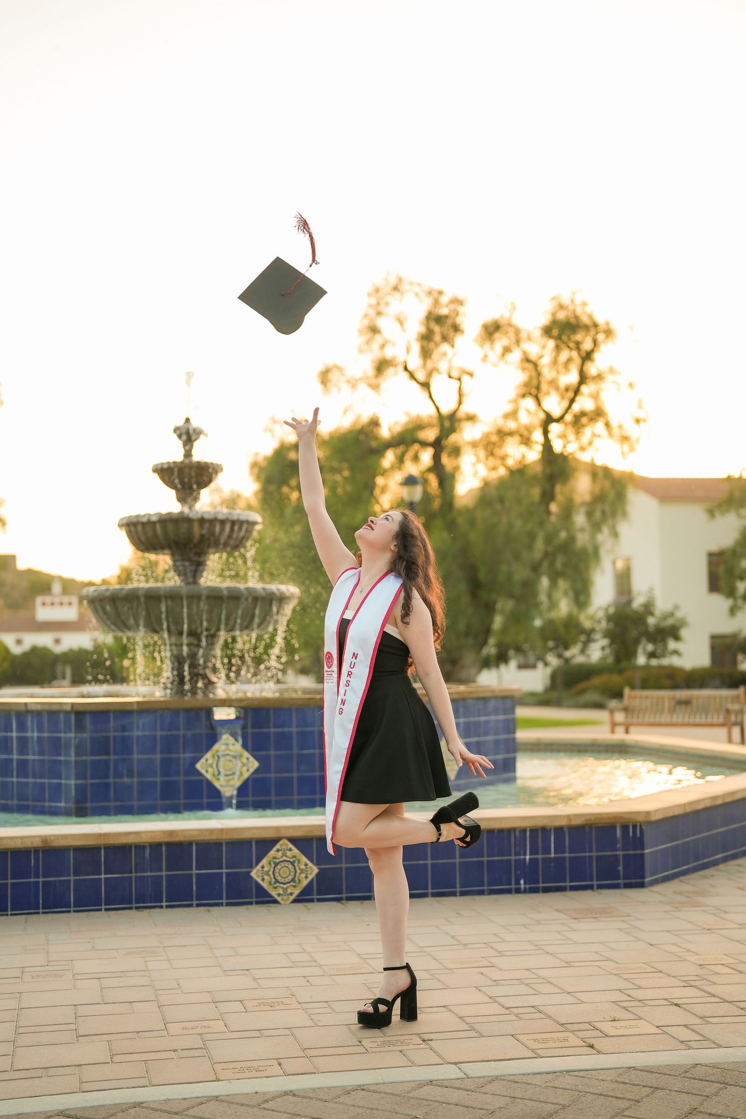 Nursing graduate in black dress and heels tosses her graduation cap into the air beside a tiled campus fountain at sunset.