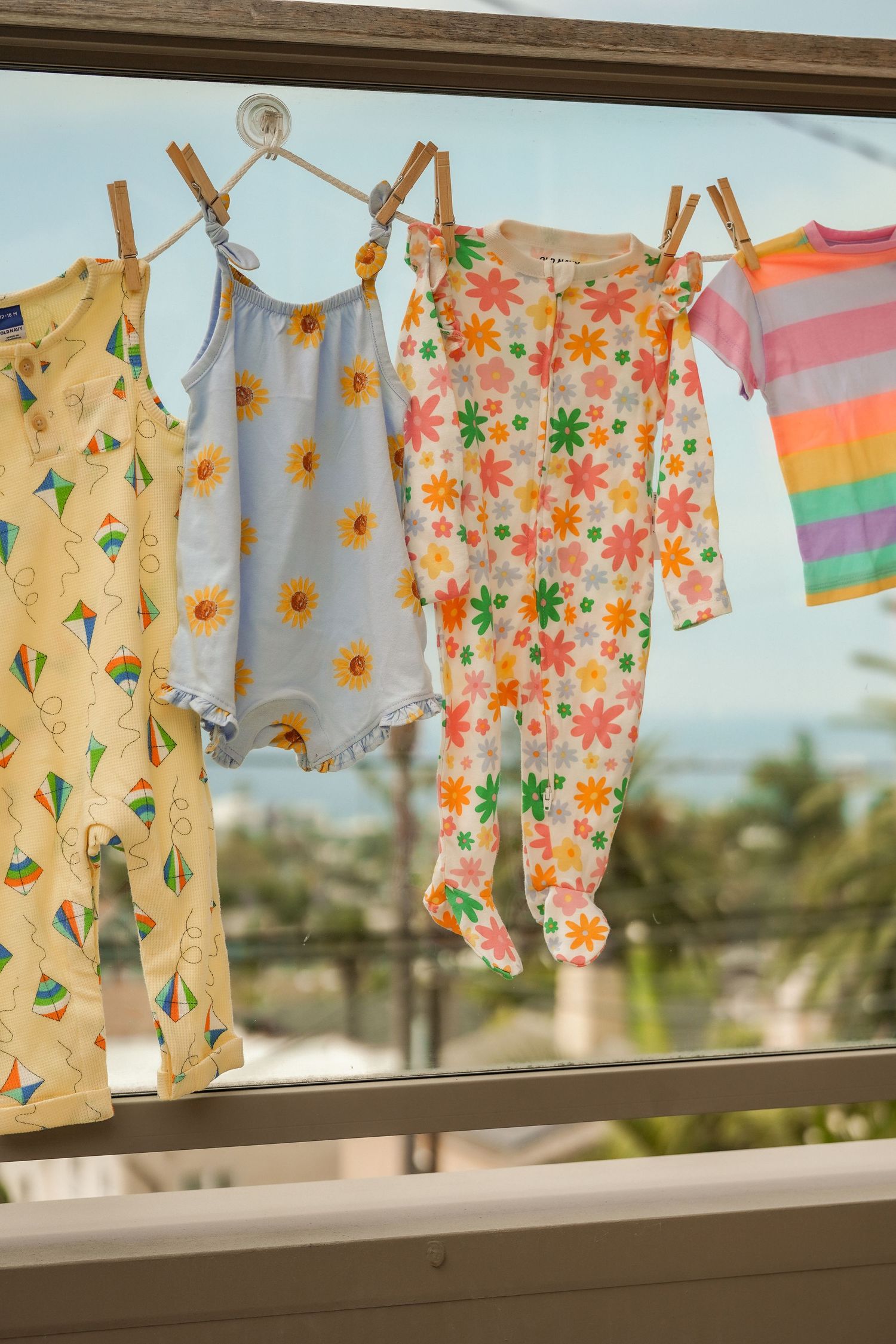 Colorful baby clothes with floral, kite and striped patterns hang on a clothesline with a sunny coastal view behind.