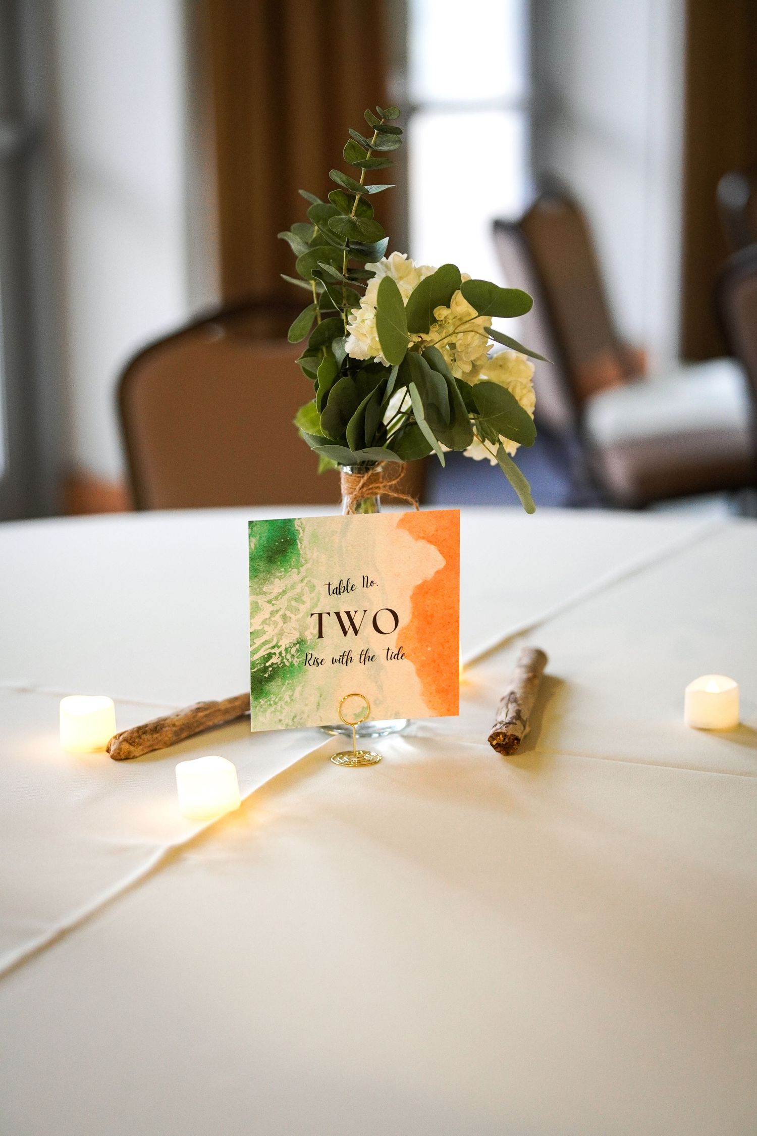 A wedding-style table centerpiece with white flowers and greenery beside a green and orange Table Two card and flameless candles.