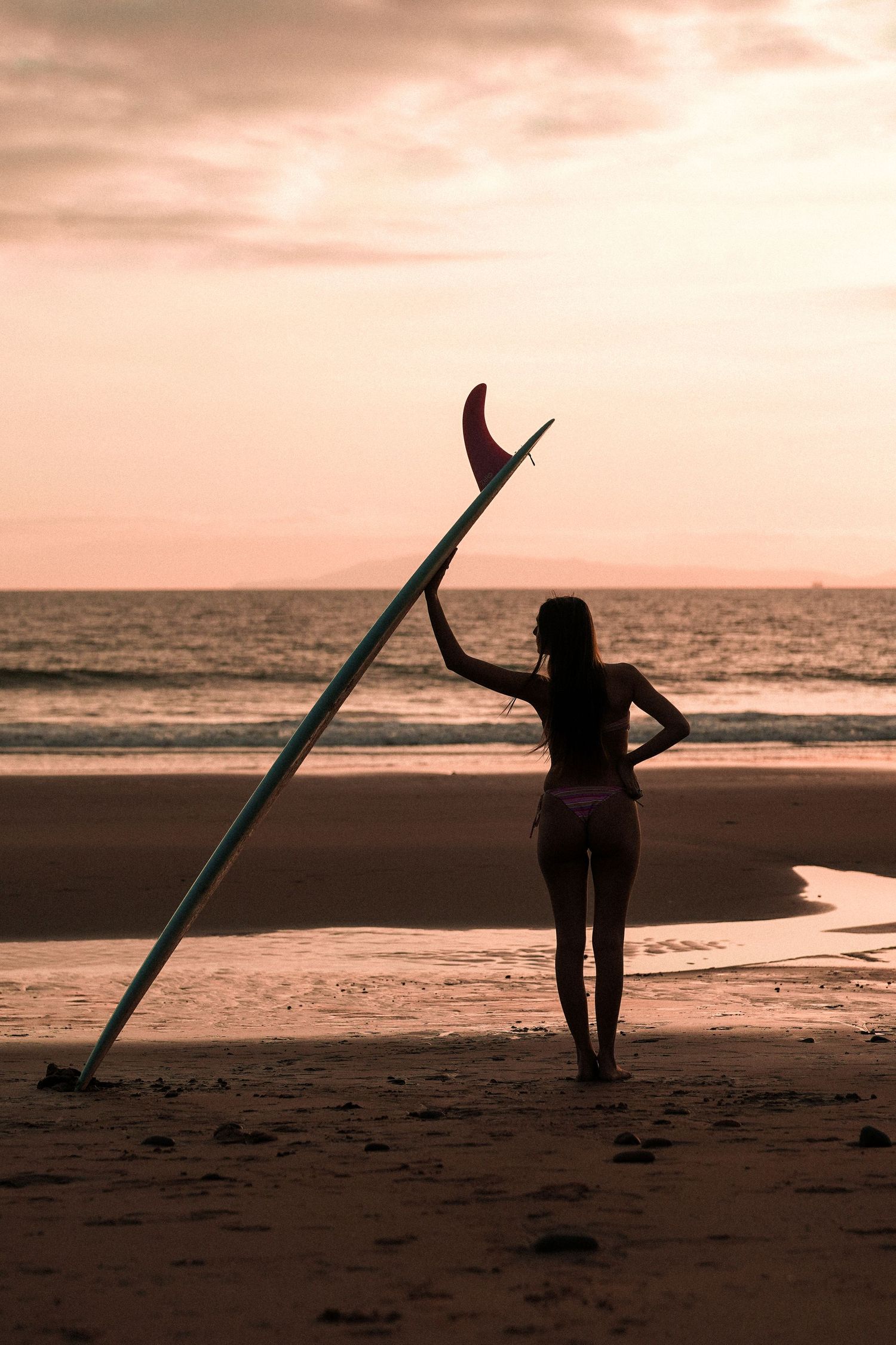 Silhouette of a woman leaning a surfboard upright on the beach as the ocean glows under a pink sunset sky.