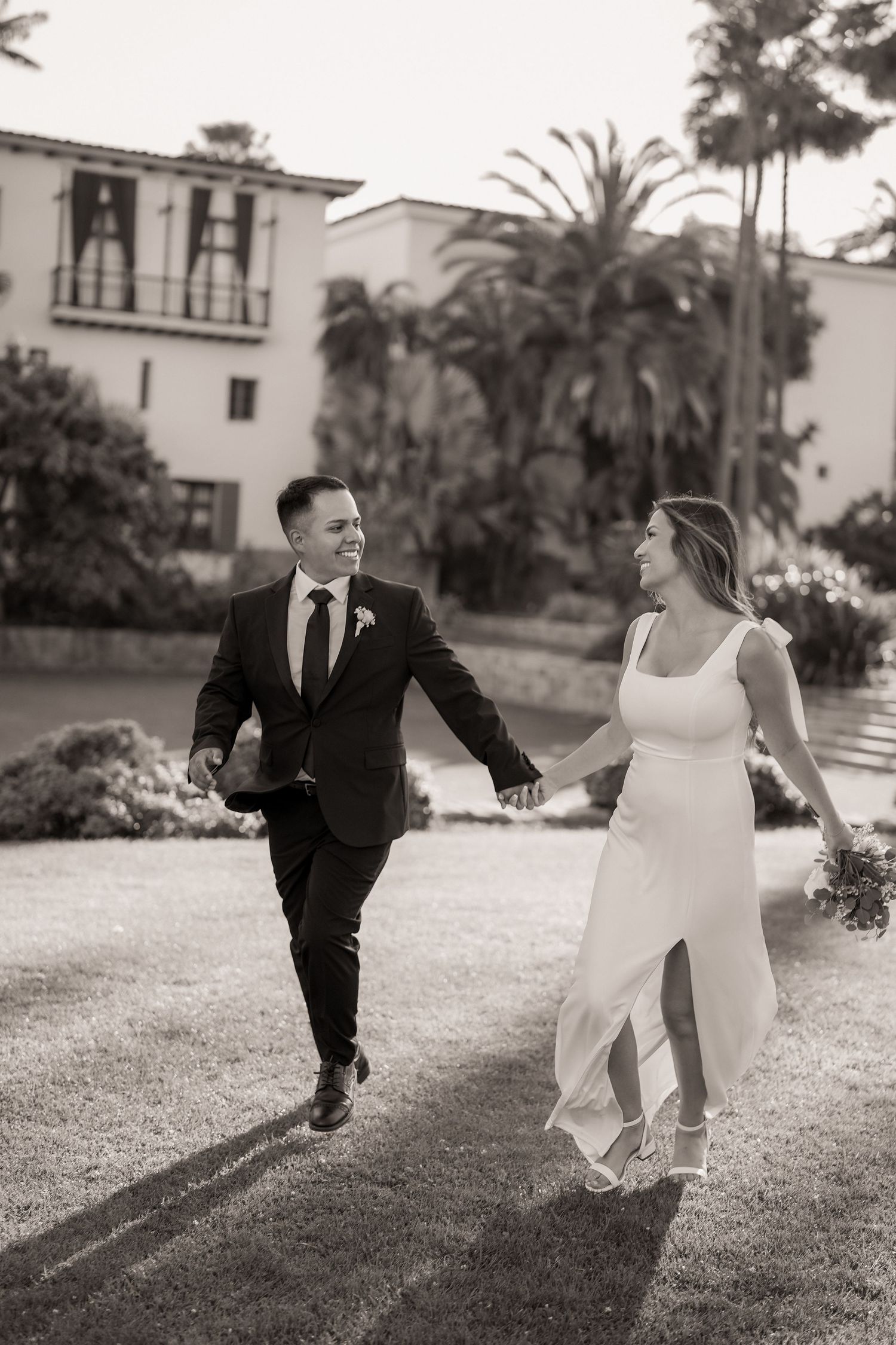 Black and white photo of a happy couple in wedding attire holding hands and running across a lawn.