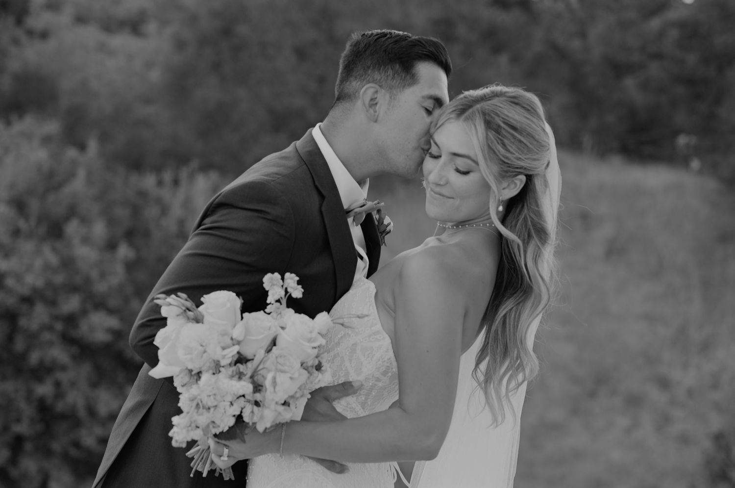 Black and white photo of a groom kissing his bride on the cheek as she holds a bouquet of flowers.