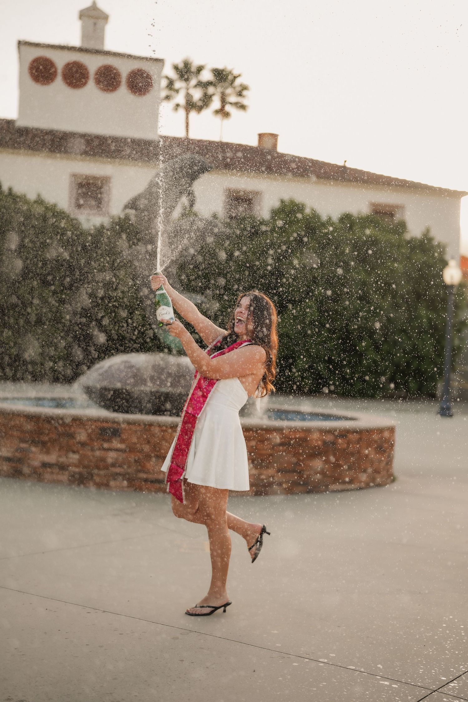 A graduate in a white dress and red sash celebrates with a spraying champagne bottle in front of a brick fountain and hedges.