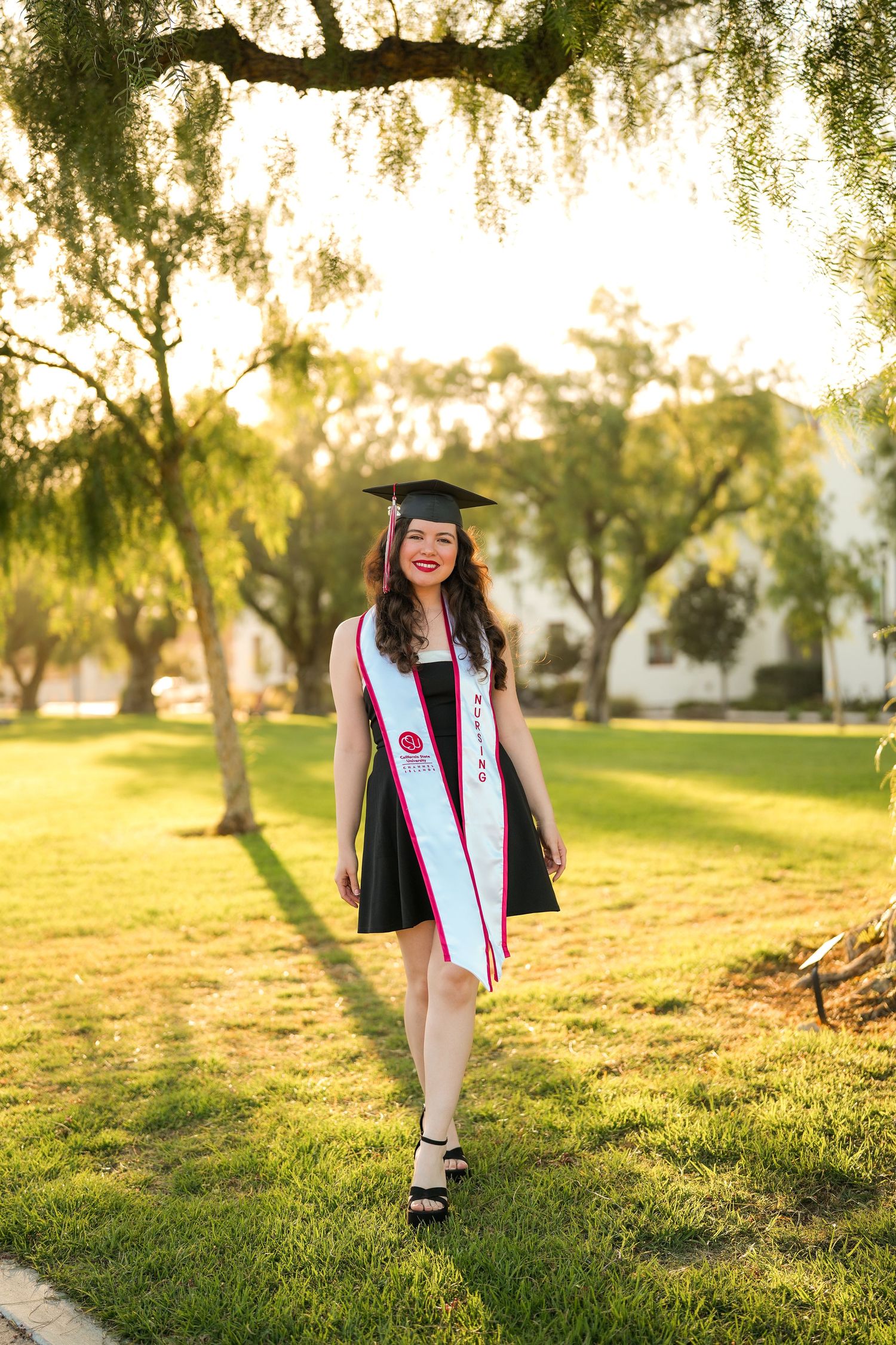 A nursing graduate in a black dress, cap, and white stole stands on a sunny grass lawn beneath trees at golden hour.