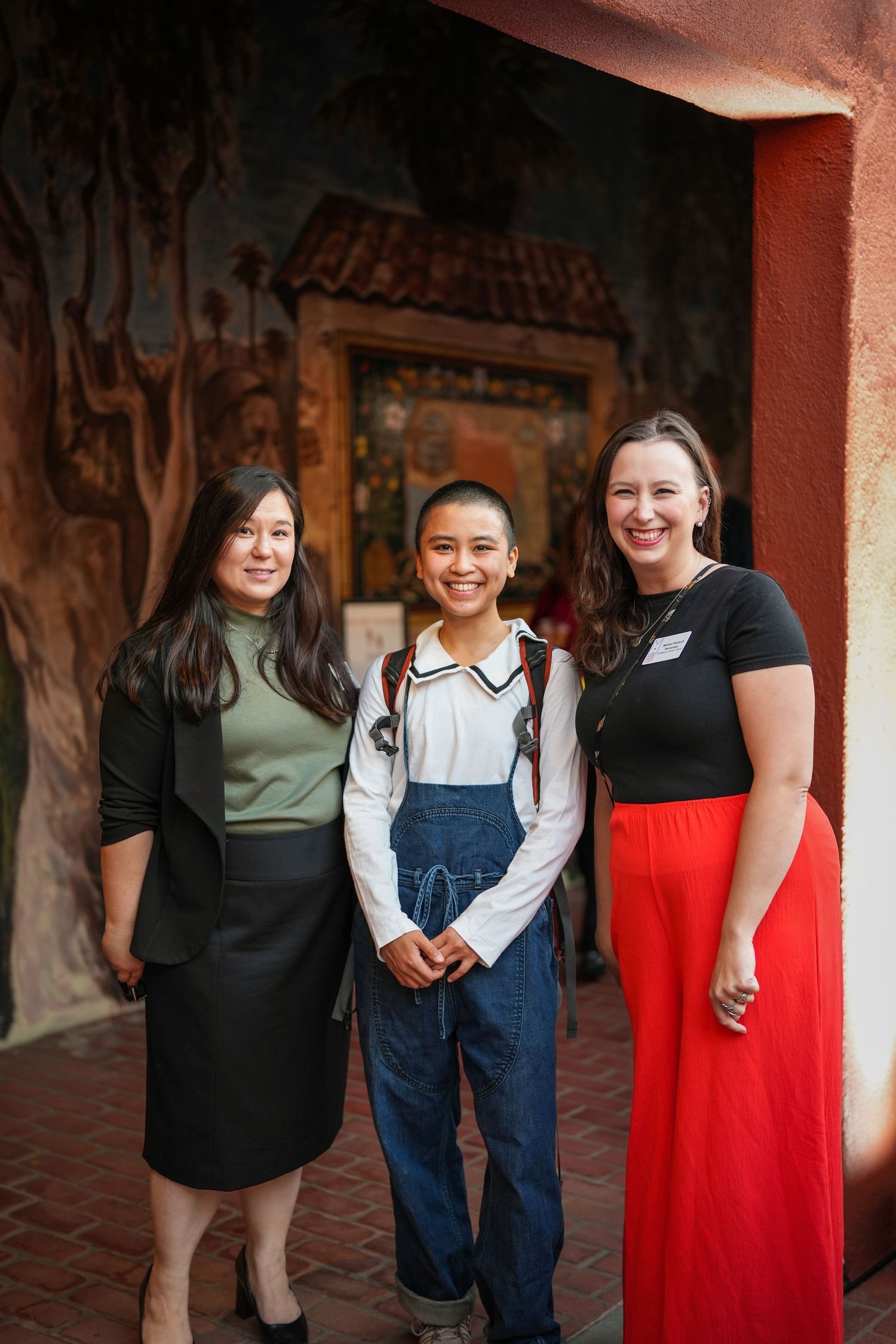 Three smiling women pose together in front of a painted mural at an evening event, two wearing name tags.