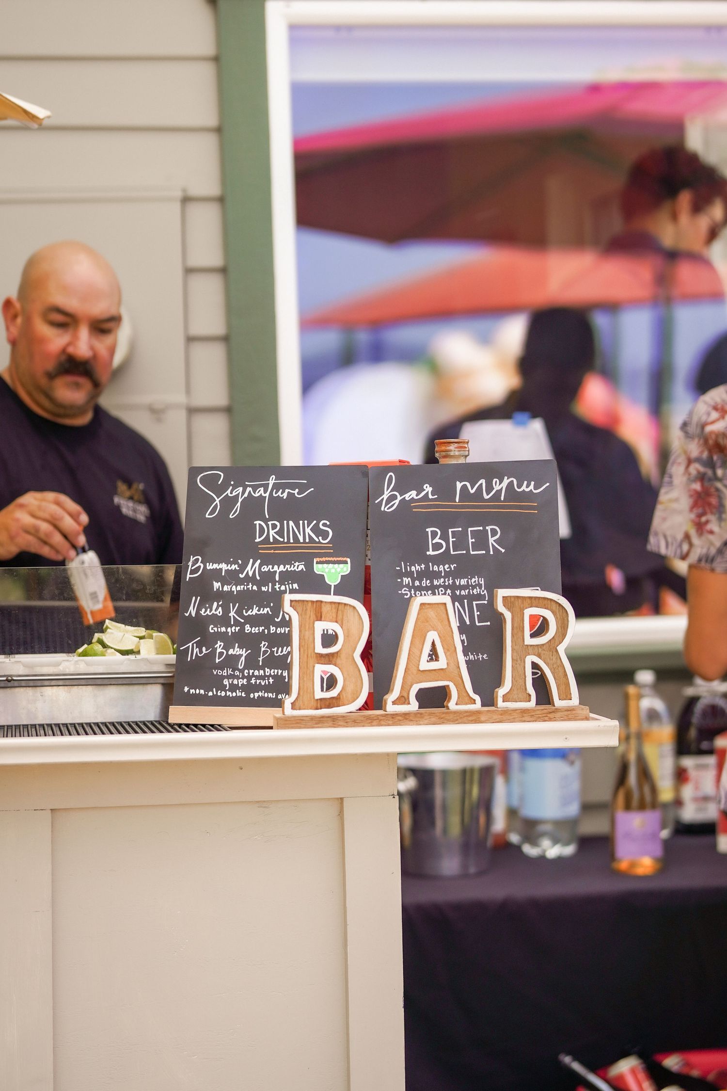 Outdoor event bar with chalkboard signature drinks and beer menu signs, wooden BAR letters, and a bartender preparing garnishes.