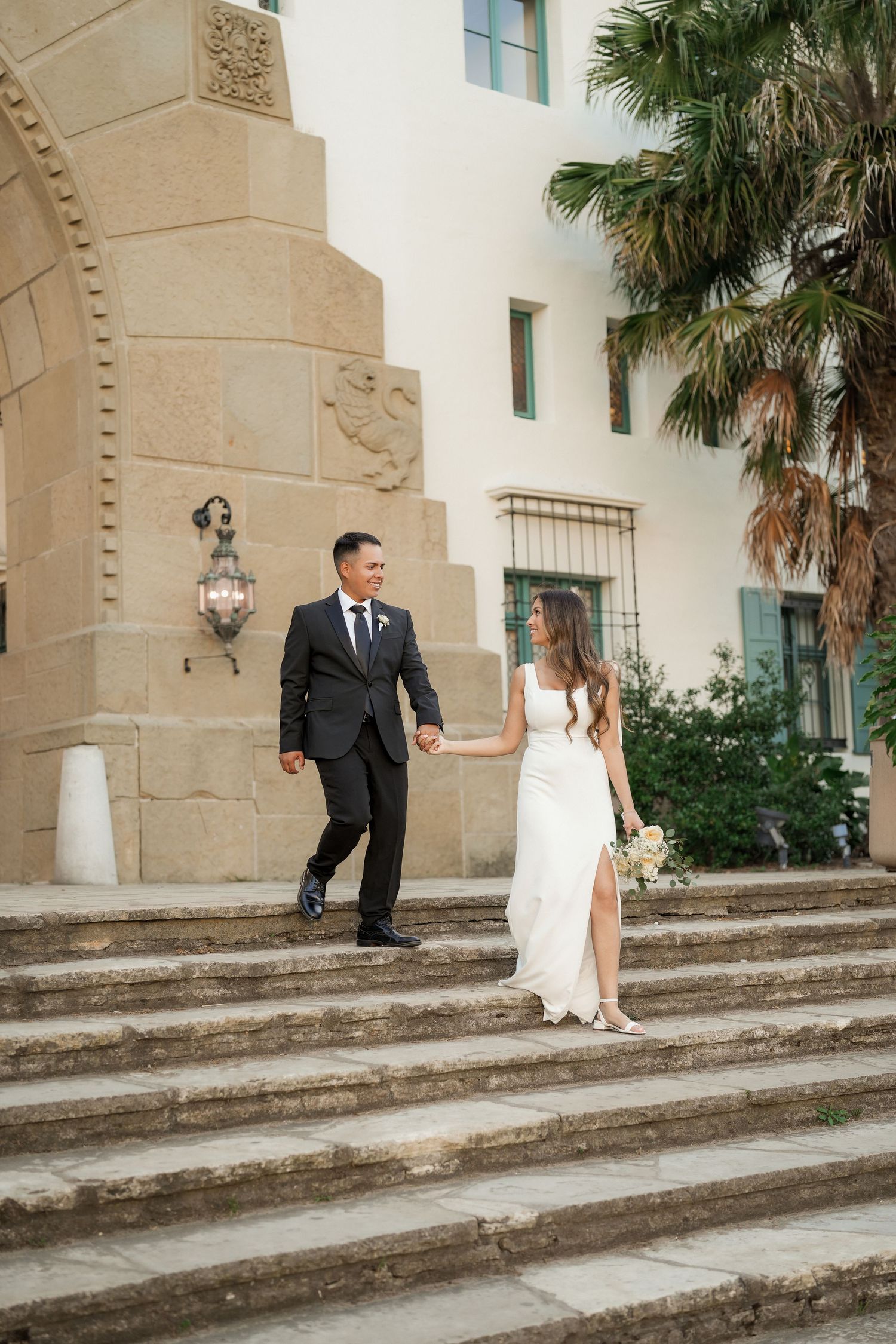 Bride and groom hold hands and walk down stone steps beside a sandstone archway and palm tree on their wedding day.