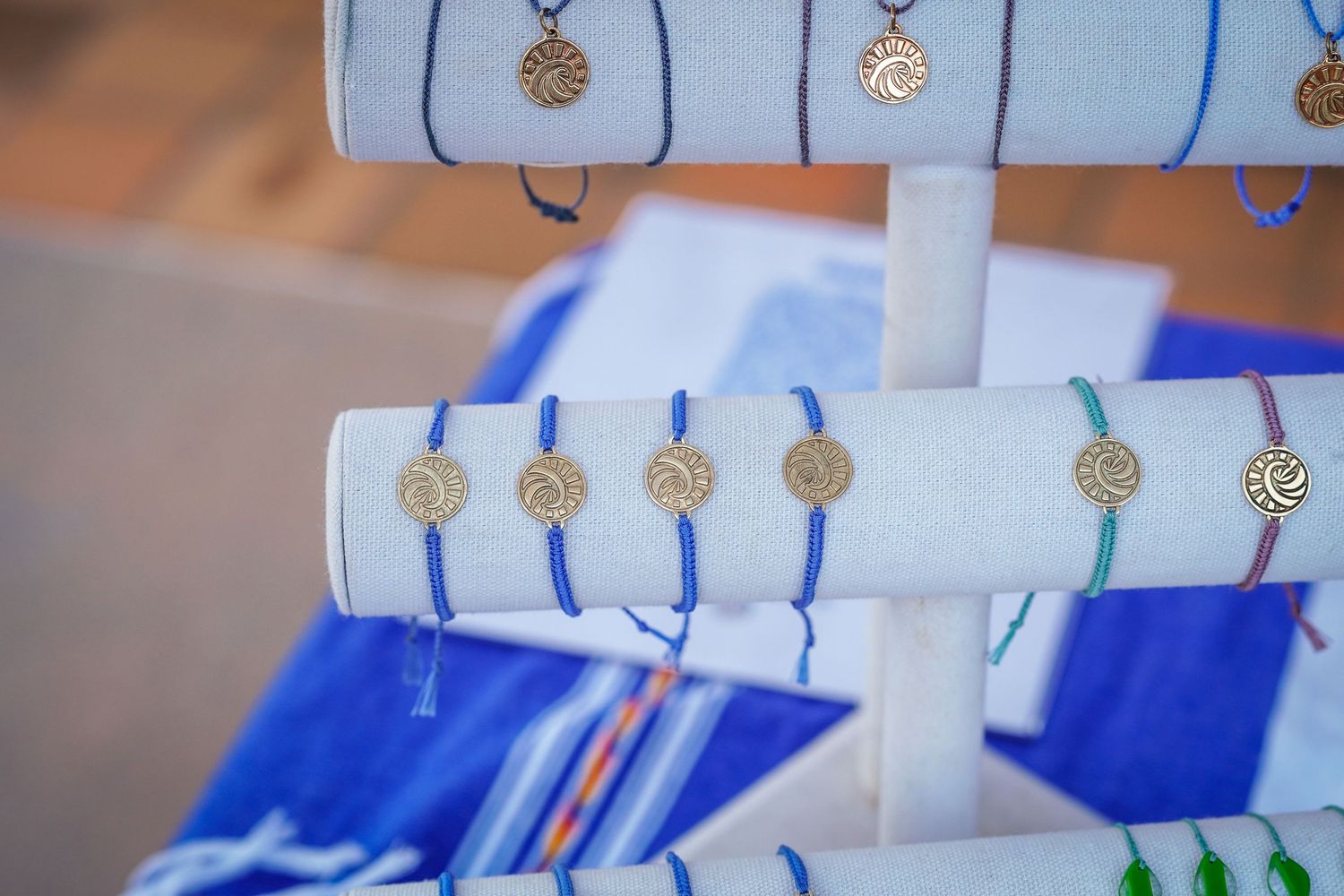 Gold sun-medallion charm bracelets with blue and teal cords displayed on white stands at a craft market booth.