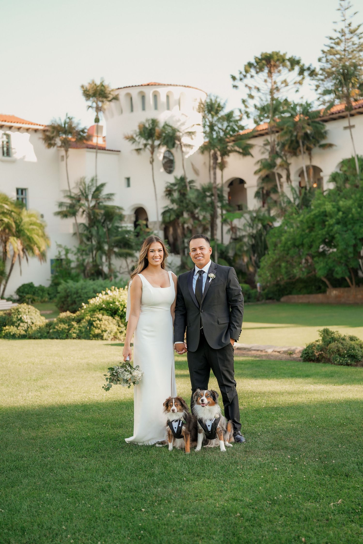 Newlyweds hold hands with two dogs in tiny bow ties at their feet on a manicured lawn before a white Spanish Colonial building.