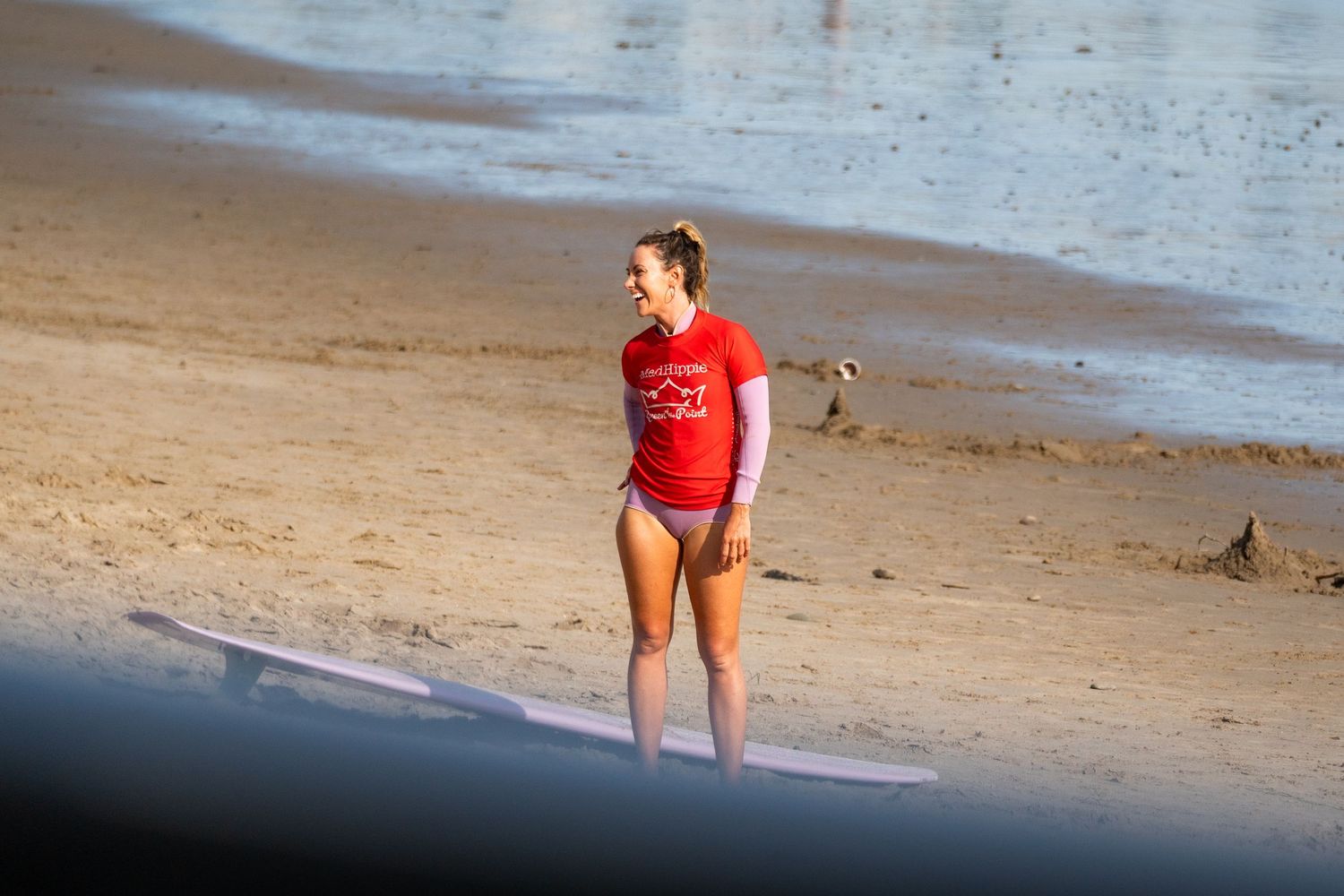 Woman in a red shirt stands on a sandy beach beside a longboard in warm evening light near the water's edge.