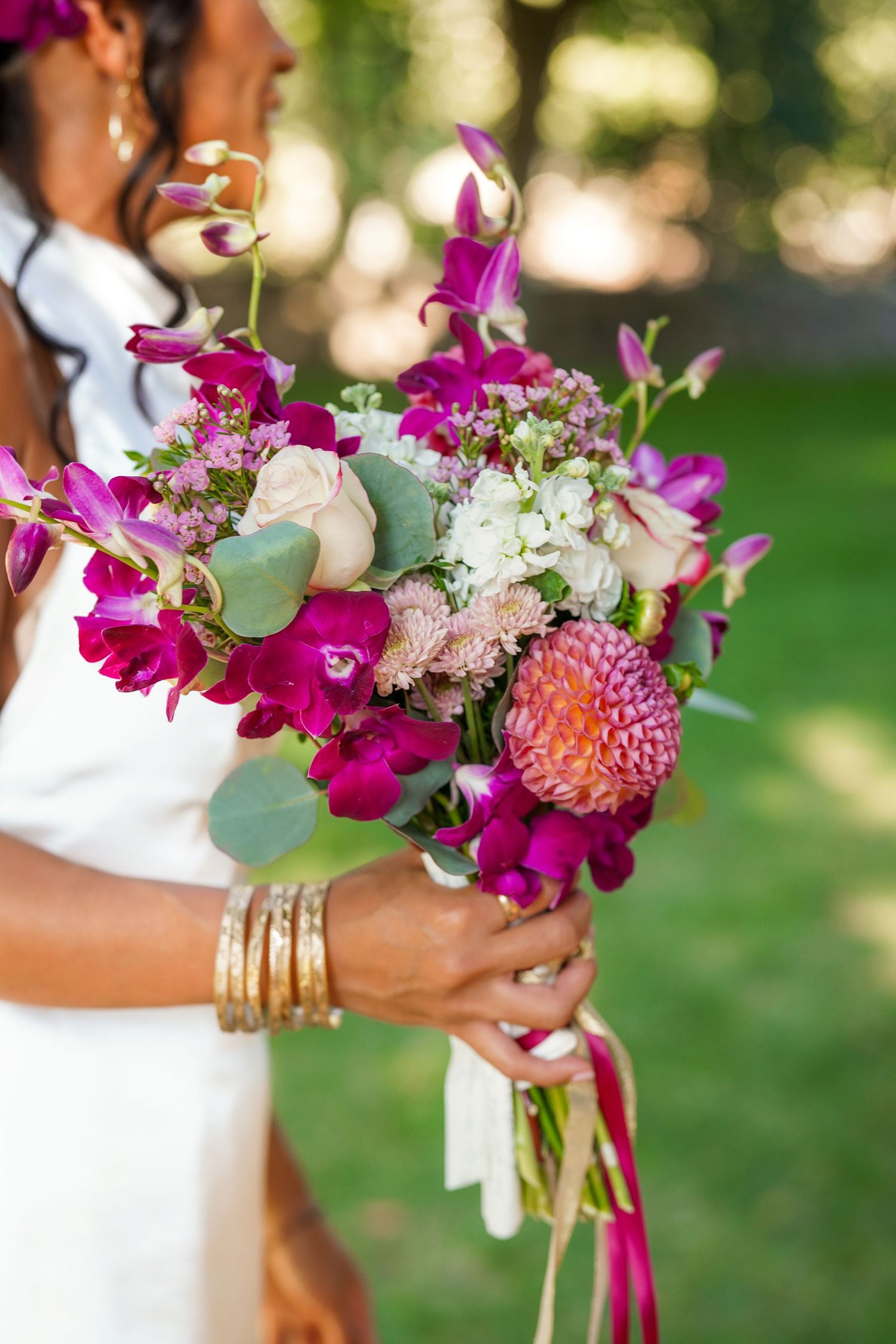 Bride in a white dress holds a vibrant bouquet of magenta orchids, pink dahlias, and white blooms, wearing gold bangles.