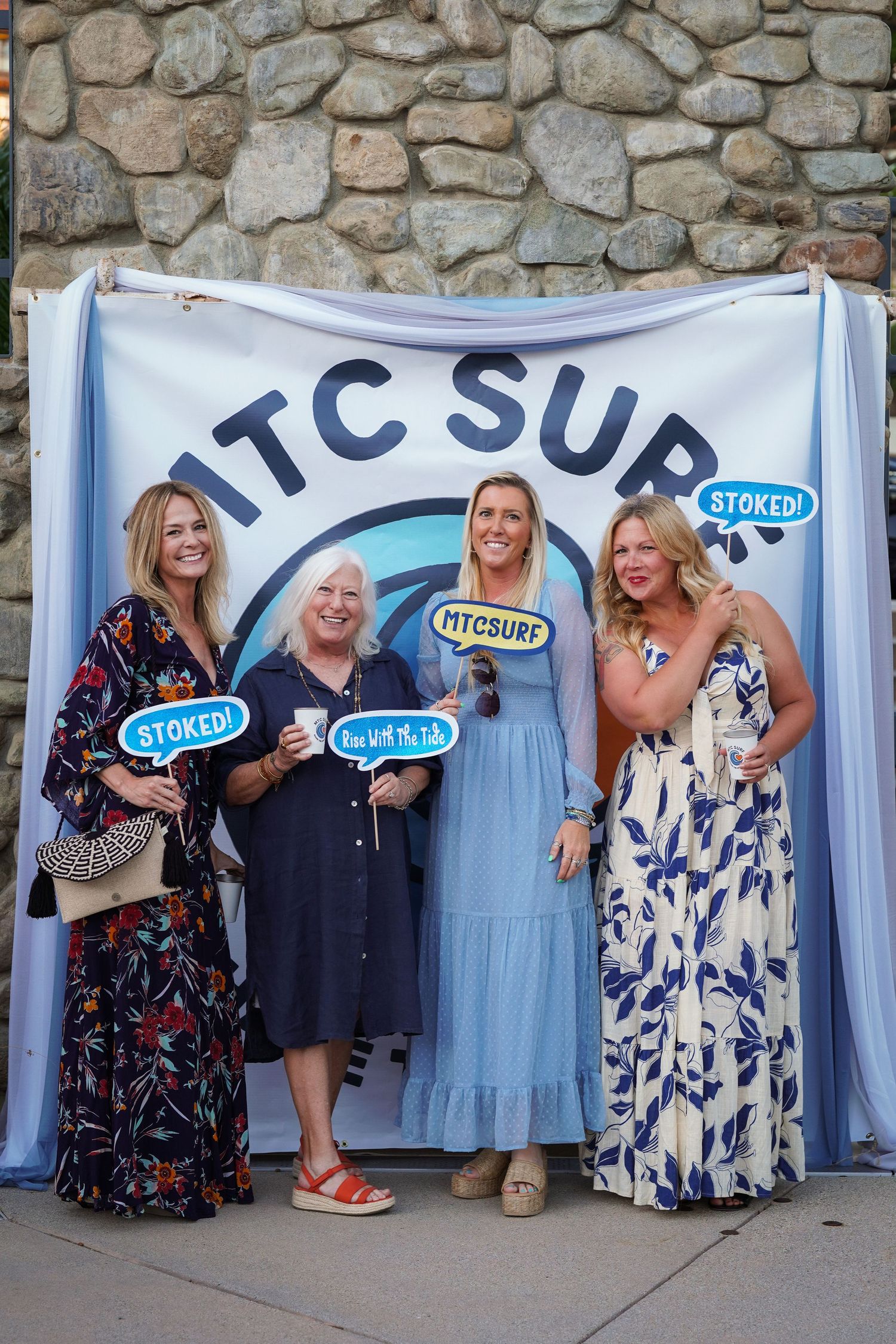 Four smiling women pose with Stoked photo booth props in front of an MTC Surf banner on a stone wall.