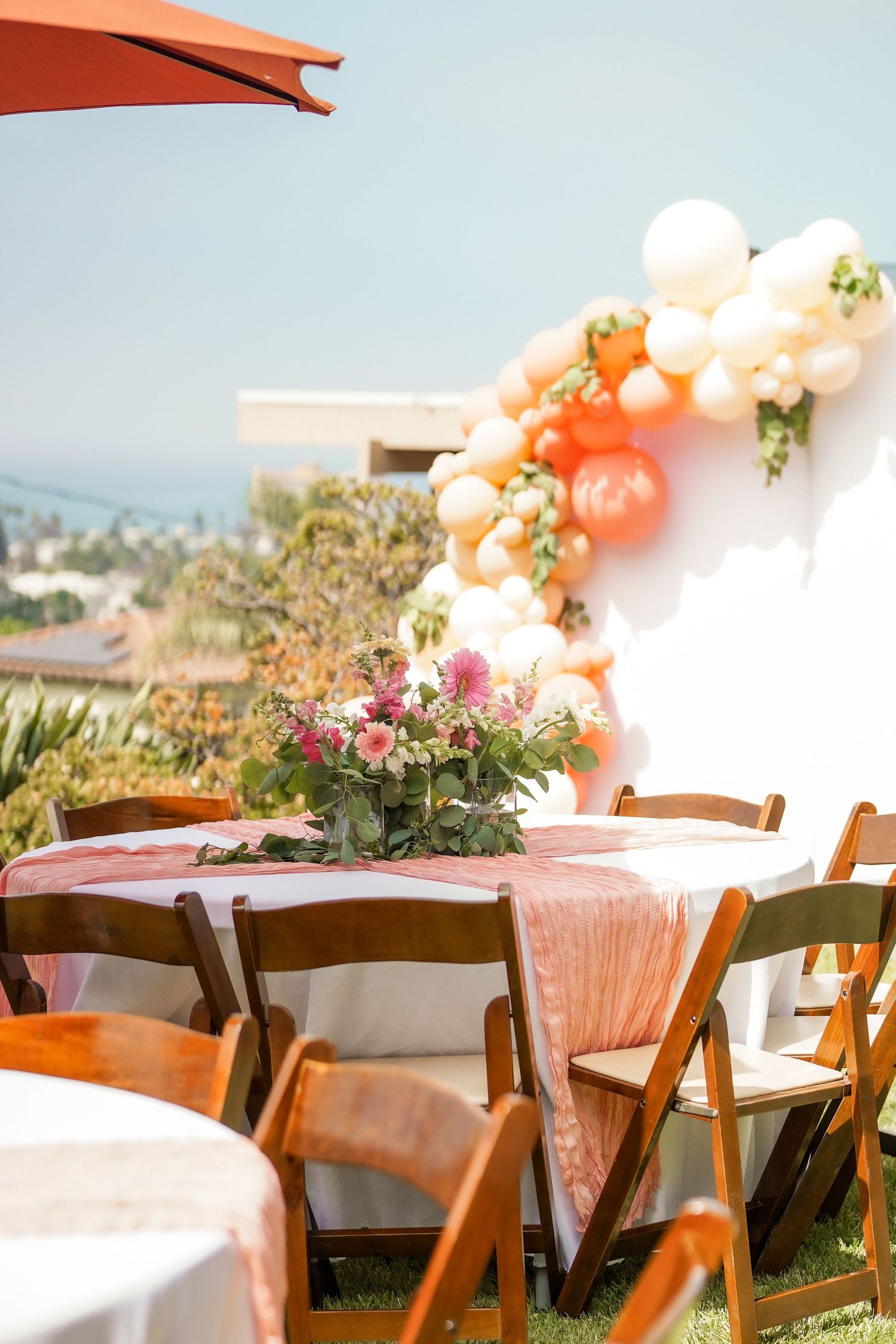 Outdoor party table with wooden folding chairs, a pink floral centerpiece and a peach and cream balloon garland against a white wall.