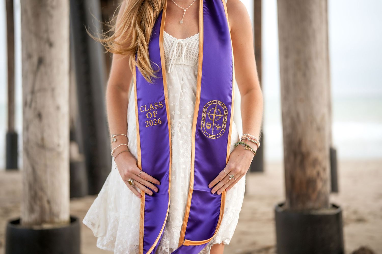 Close view of a graduate in a white dress wearing a purple Class of 2026 stole beneath a wooden pier on the beach.