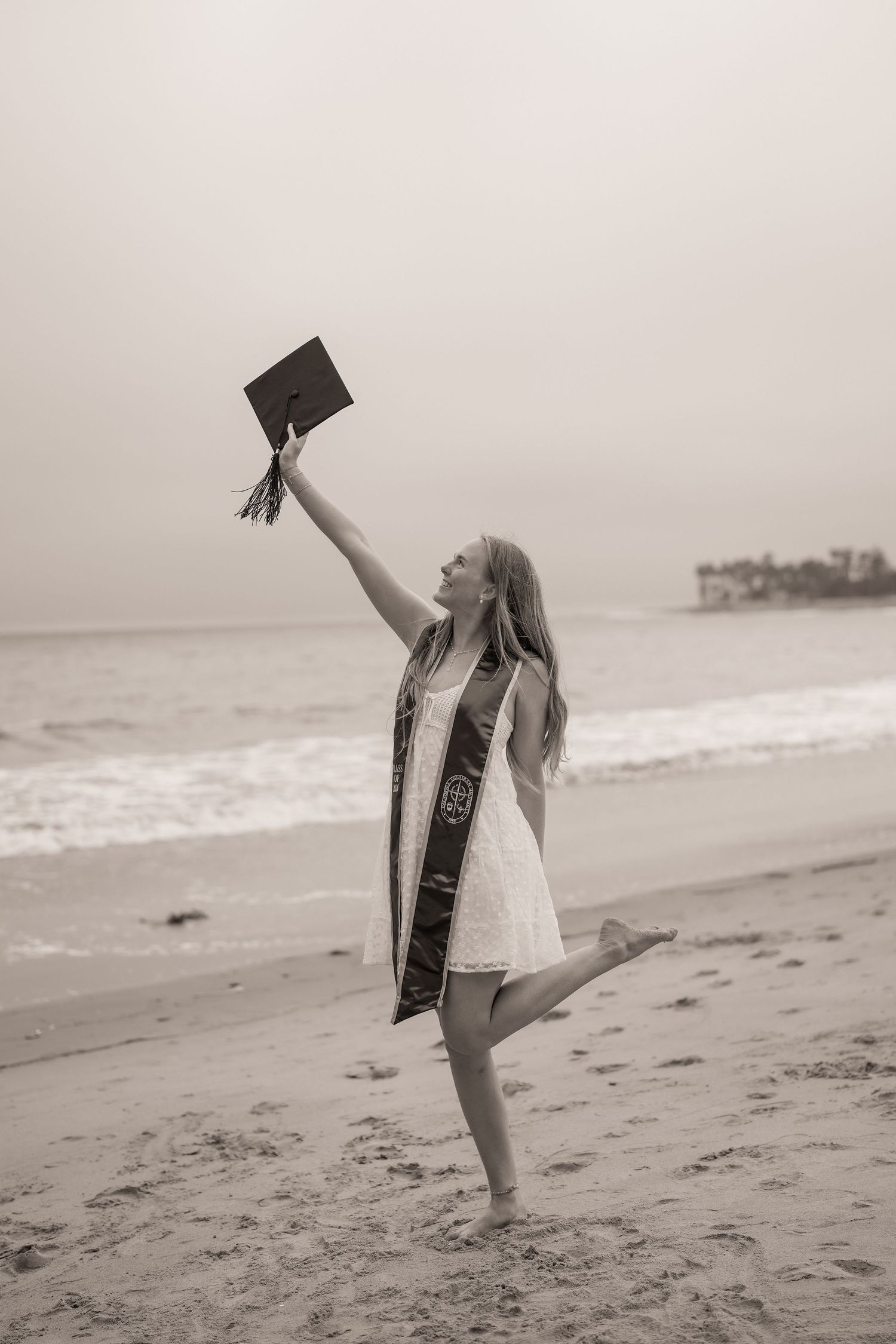 Black and white photo of a joyful graduate on one leg tossing a graduation cap into the air on the beach.