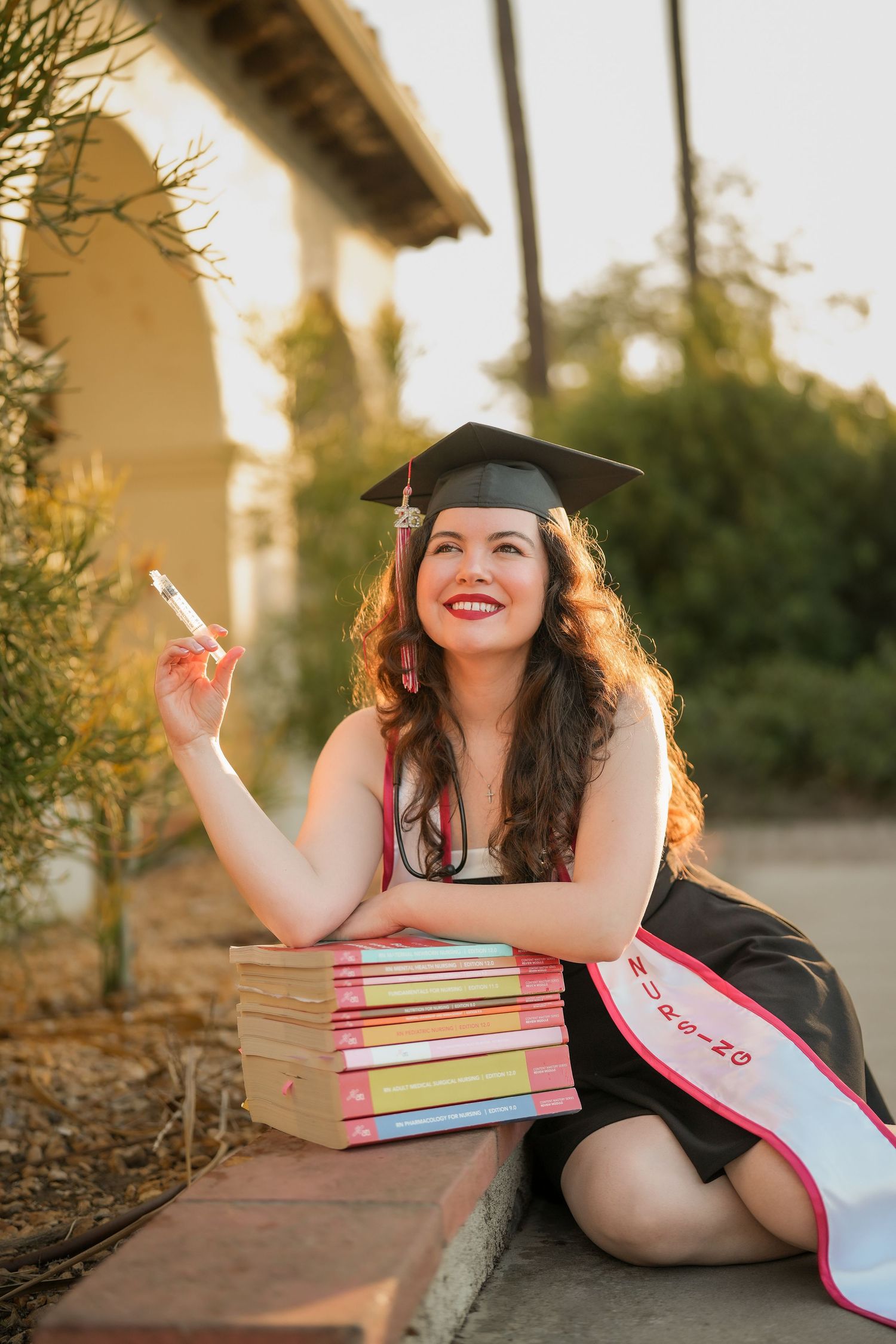Joyful nursing graduate in cap and pink-trimmed sash looks up with a smile while posing with stacked books near a Spanish-style archway.
