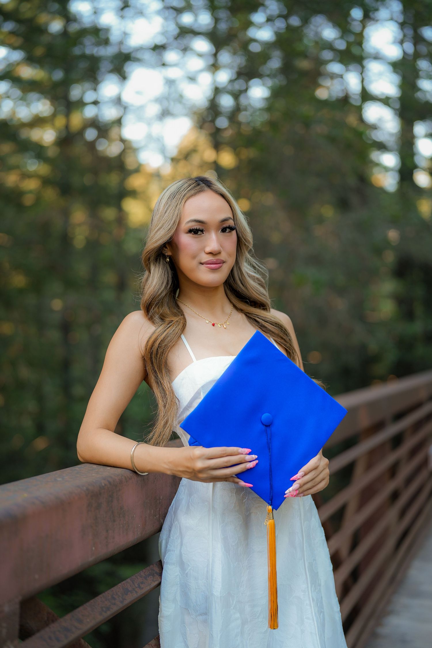 Graduate in a white dress leans on a wooden bridge railing in a sunlit forest, holding a blue cap with a gold tassel.