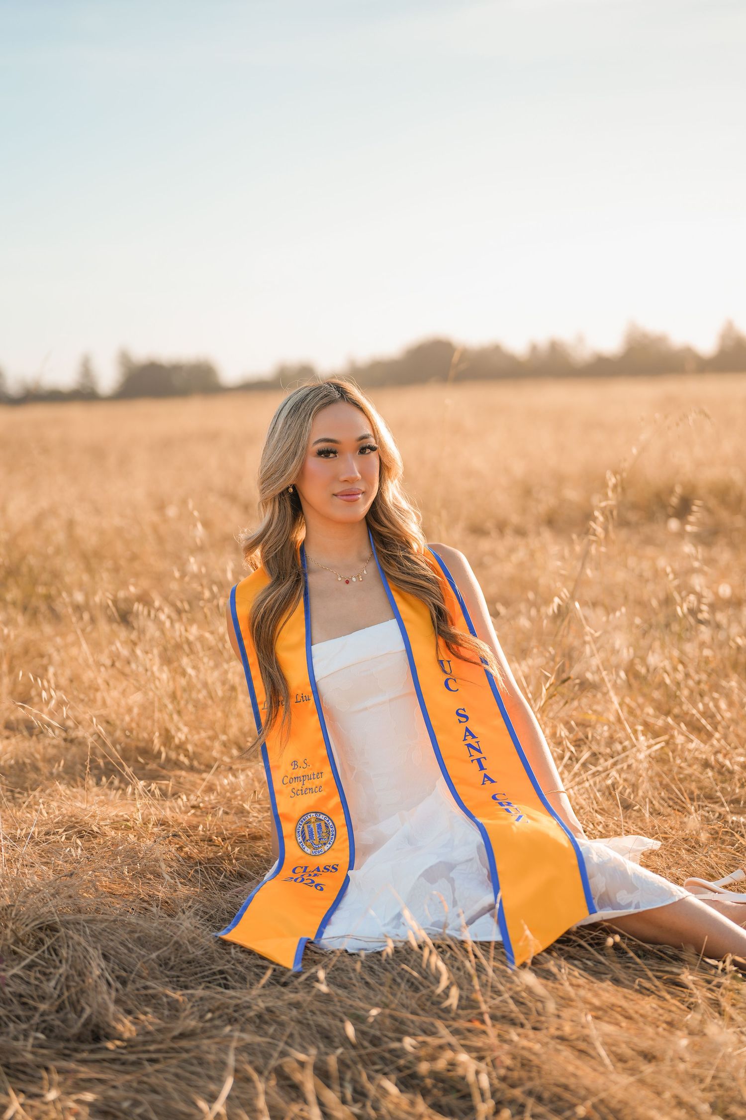 Smiling graduate in a white dress and orange UC Santa Cruz stole sits in dry golden grass during a sunset photo shoot.