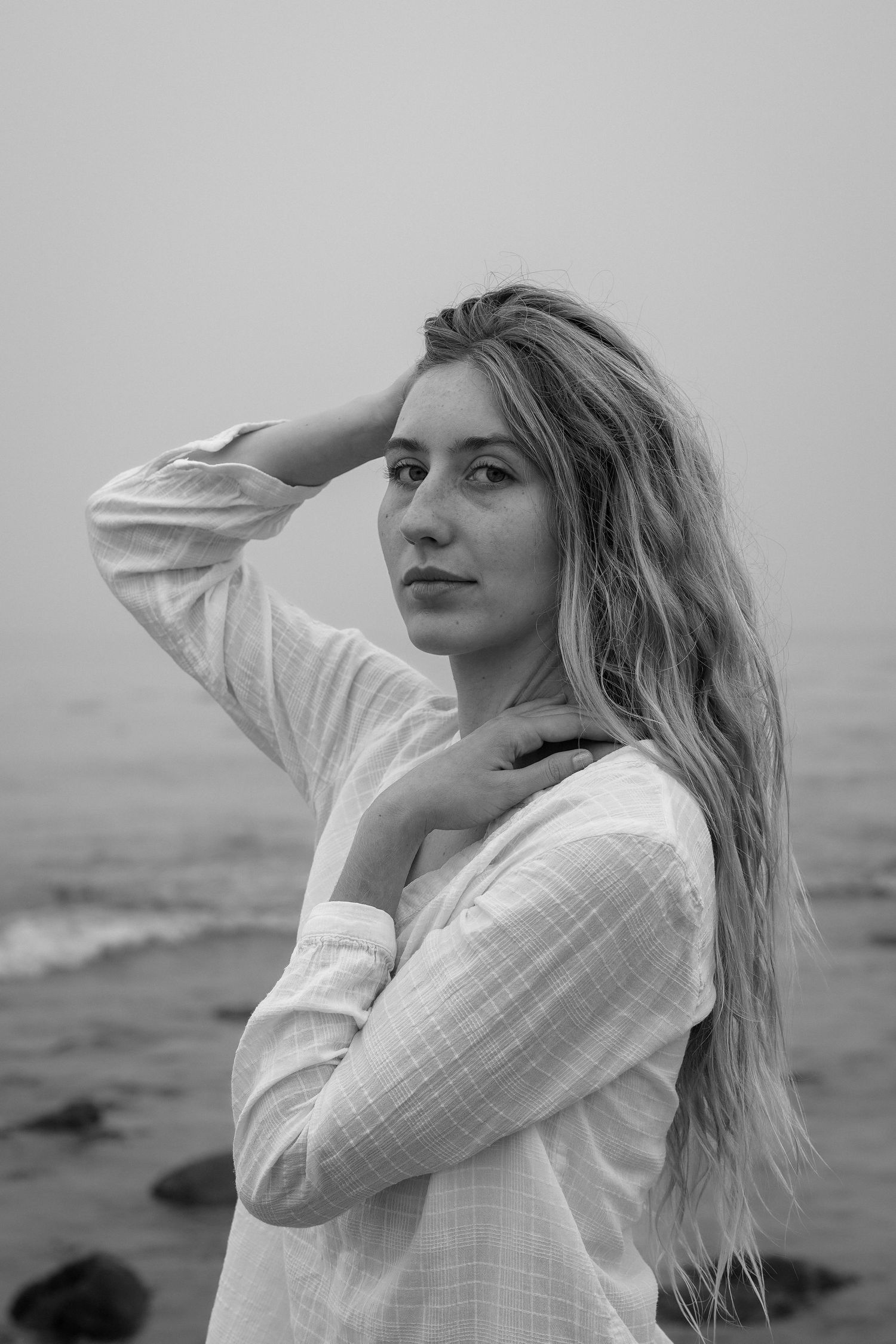Black and white portrait of a woman with long wavy hair in a white shirt, looking over her shoulder beside a misty sea.