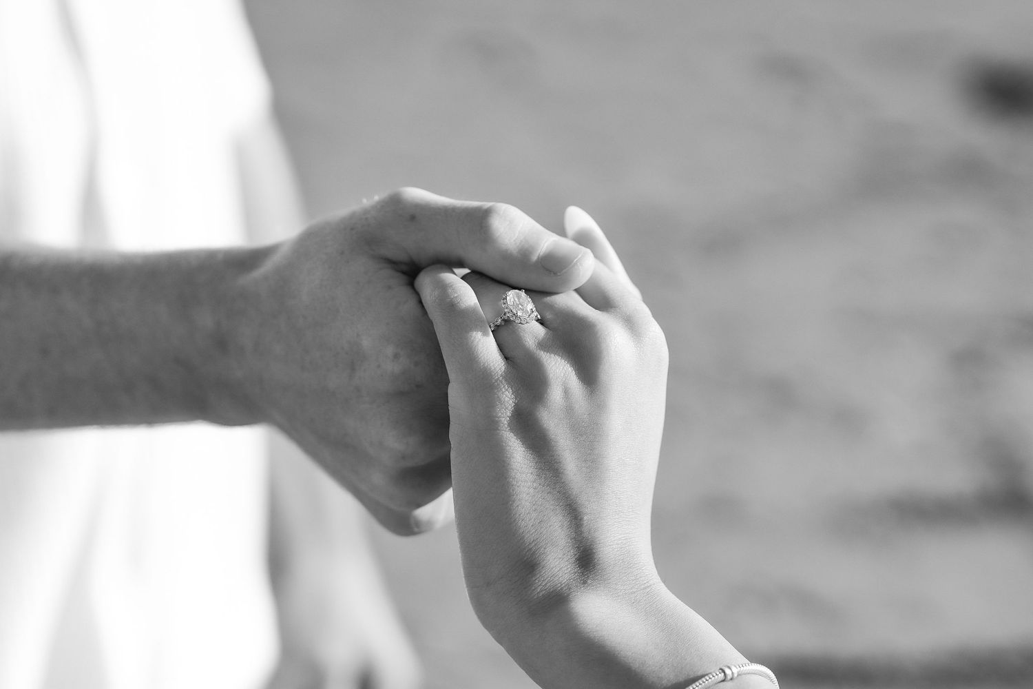 Black and white close-up of a couple's hands together on a beach, showcasing a sparkling engagement ring.