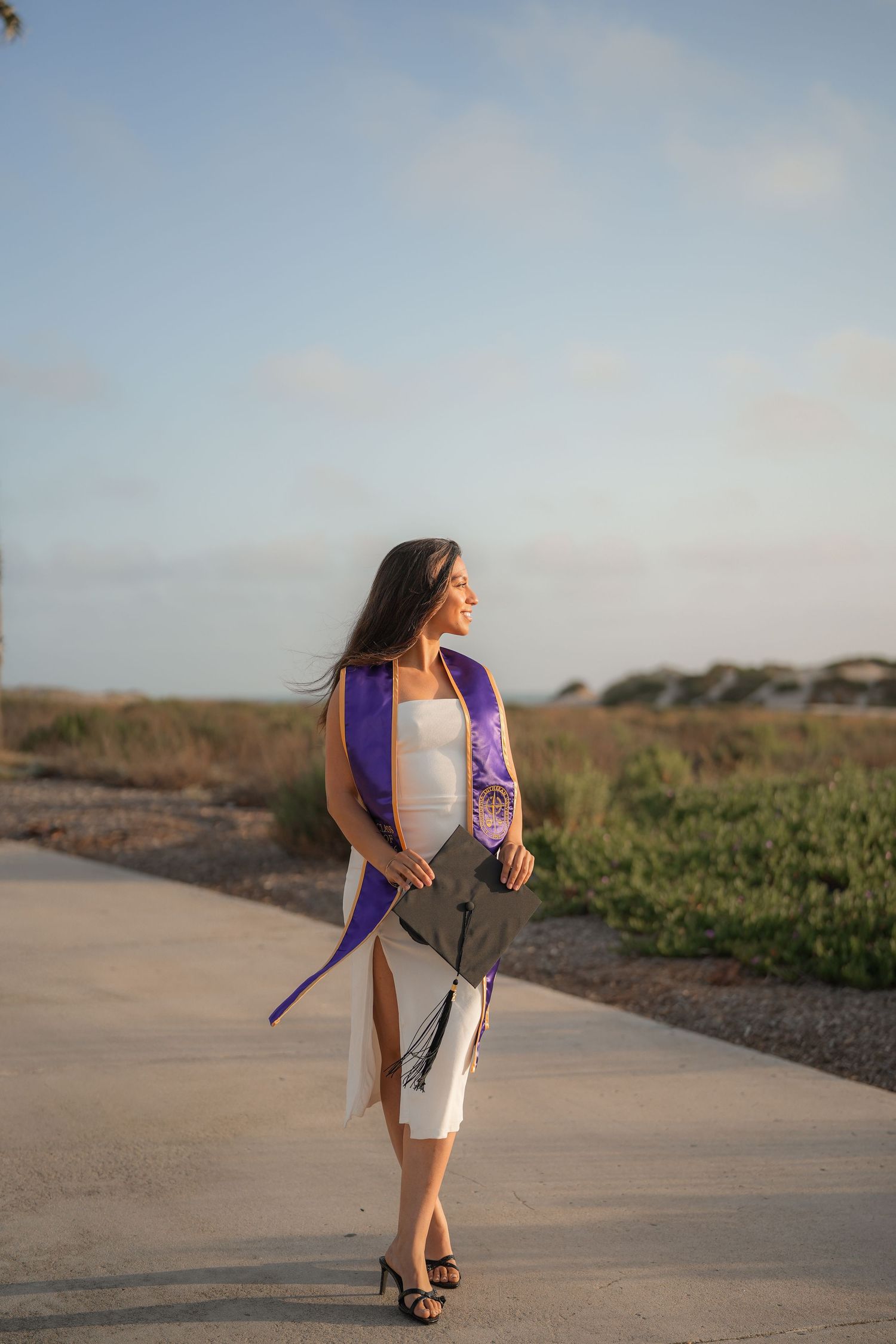 Graduate in a white dress, purple stole and black heels walks along a coastal path holding her cap at golden hour.