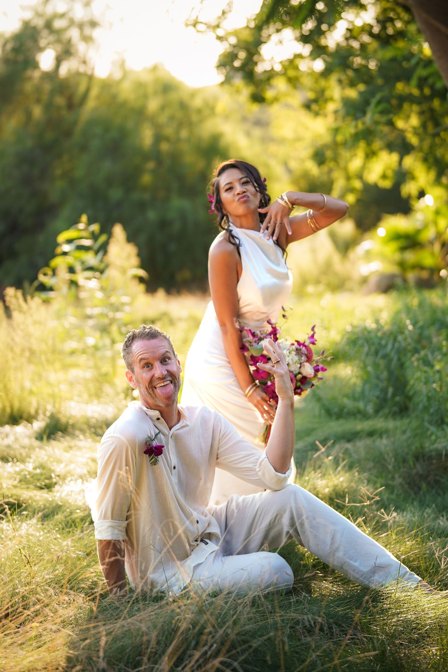 Playful groom sits in the grass making a funny face while the bride in a white gown strikes a silly pose behind him.