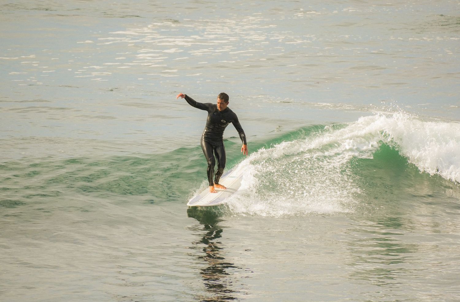 Surfer in a black wetsuit balances with arms outstretched while riding a small green wave on a longboard.