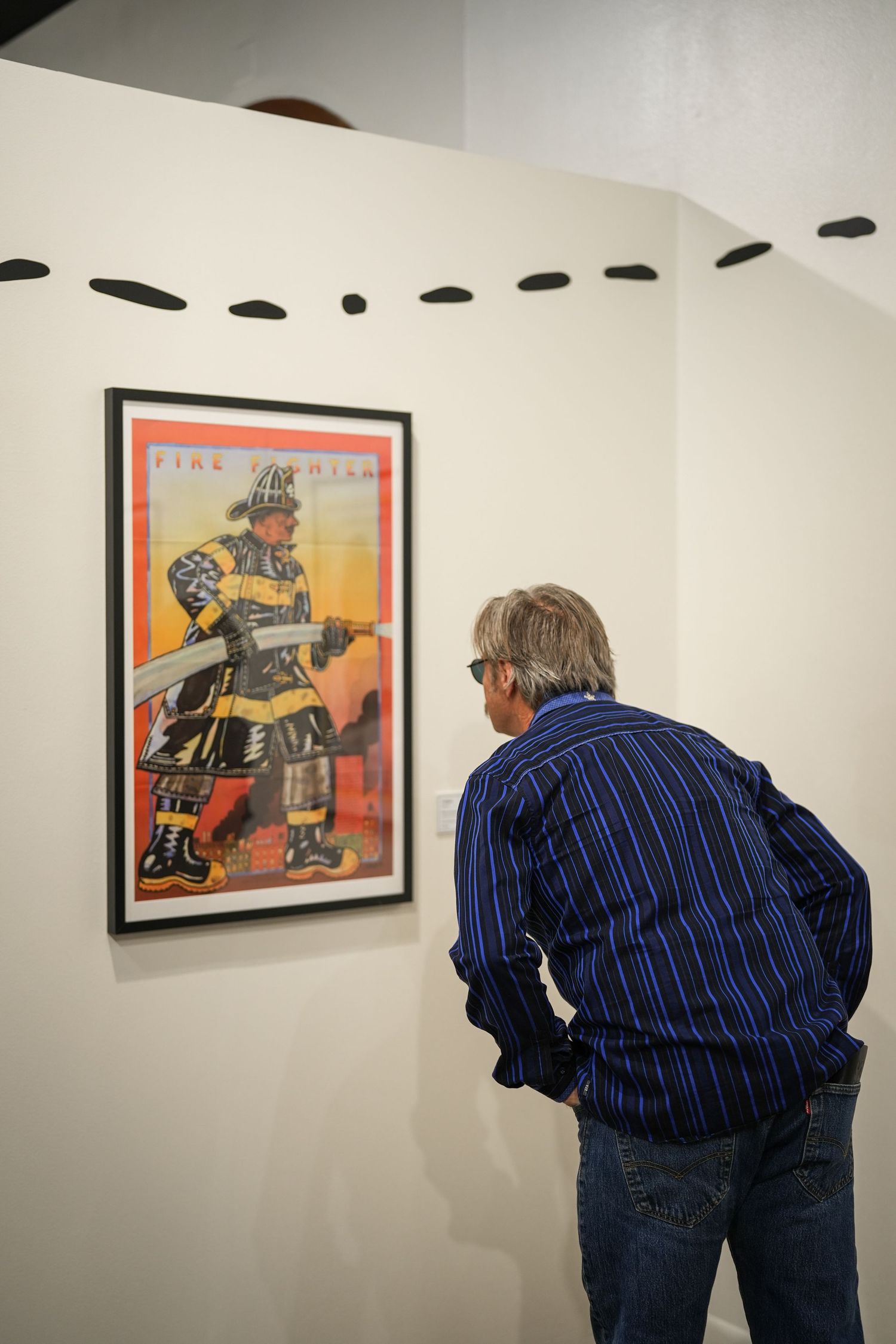 A man in a blue striped shirt leans in to examine a framed firefighter poster on a gallery wall.