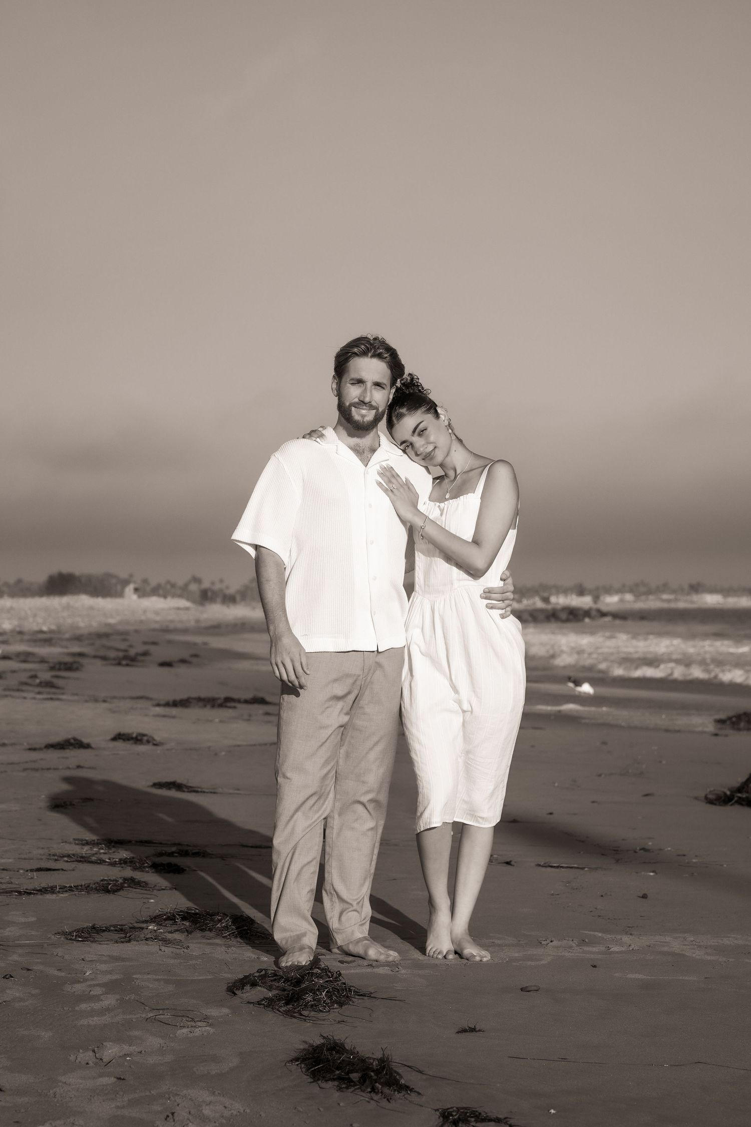 Black and white photo of a couple embracing on a beach, the woman resting her head on the man's shoulder.