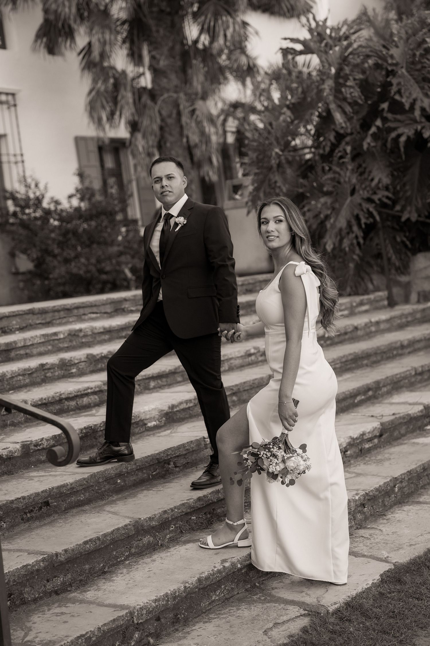 Black and white portrait of a groom and a bride holding a bouquet posing on stone steps surrounded by tropical plants.