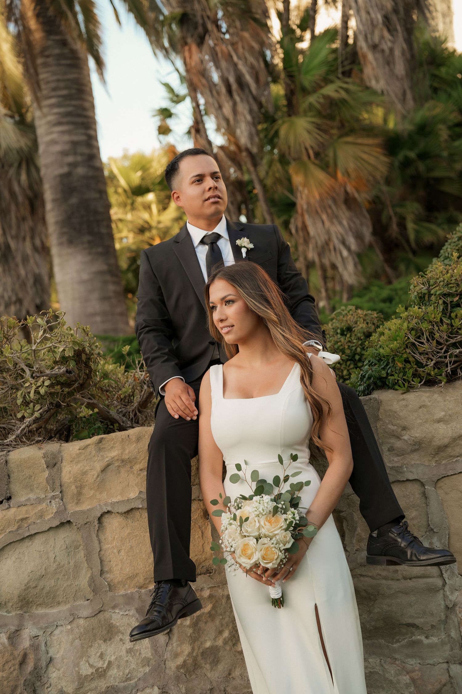 A bride in a white dress holding a bouquet stands before a groom in a black suit seated on a stone wall surrounded by palm trees.