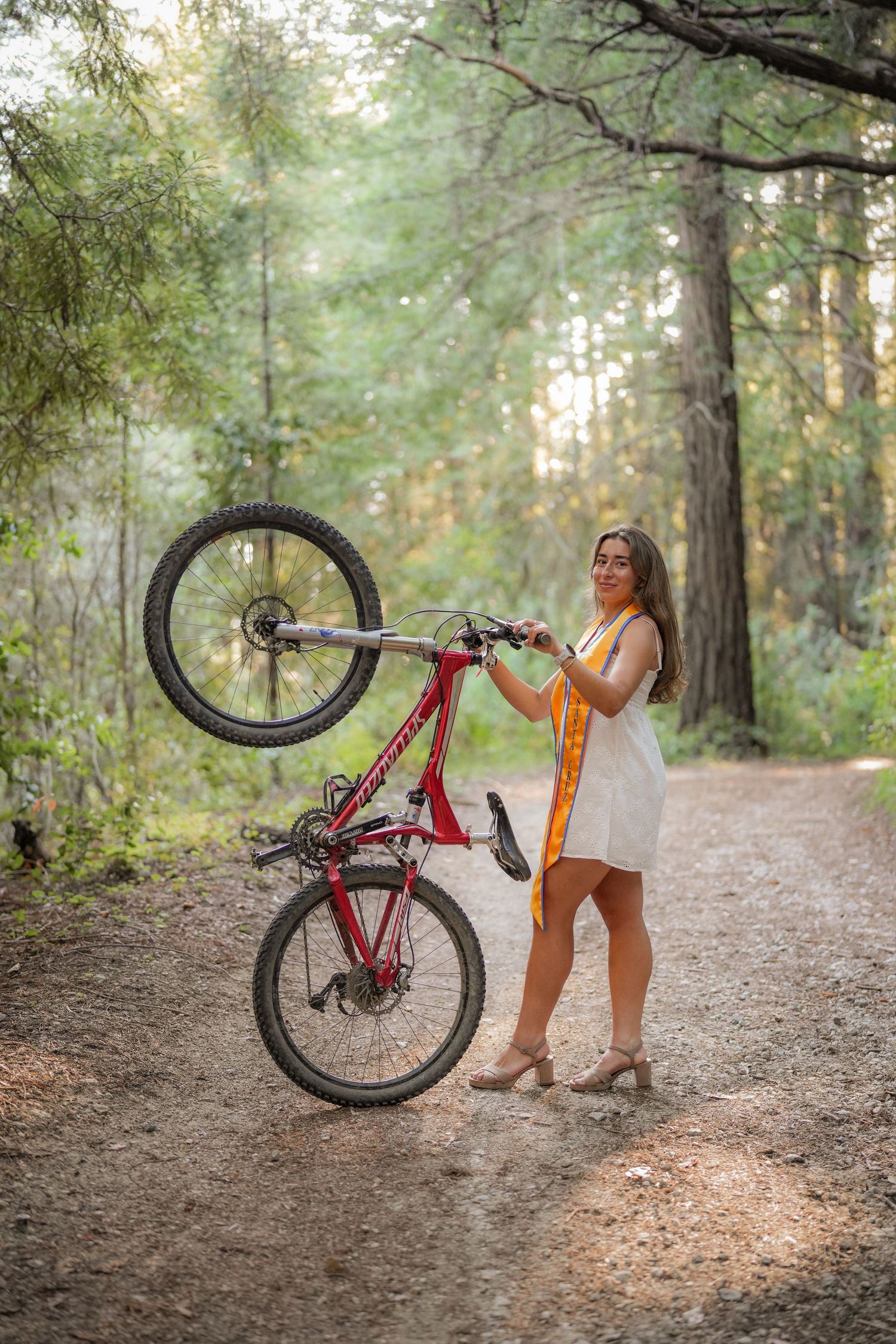 Graduate in a white dress and gold stole smiles while hoisting a red mountain bike onto its back wheel in the woods.